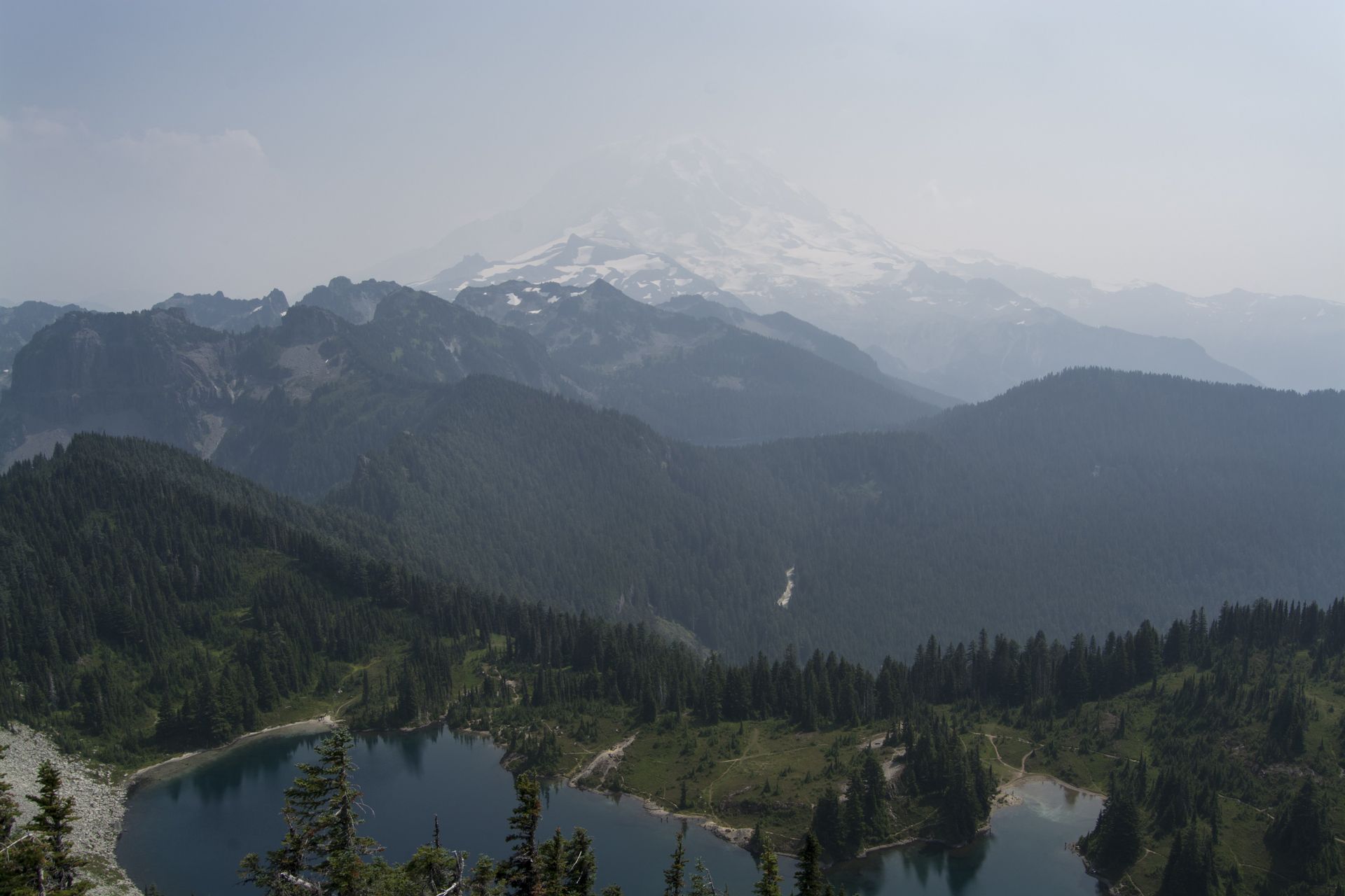 Mountain landscape with lakes, forested hills, and a snow-capped mountain under a hazy sky.