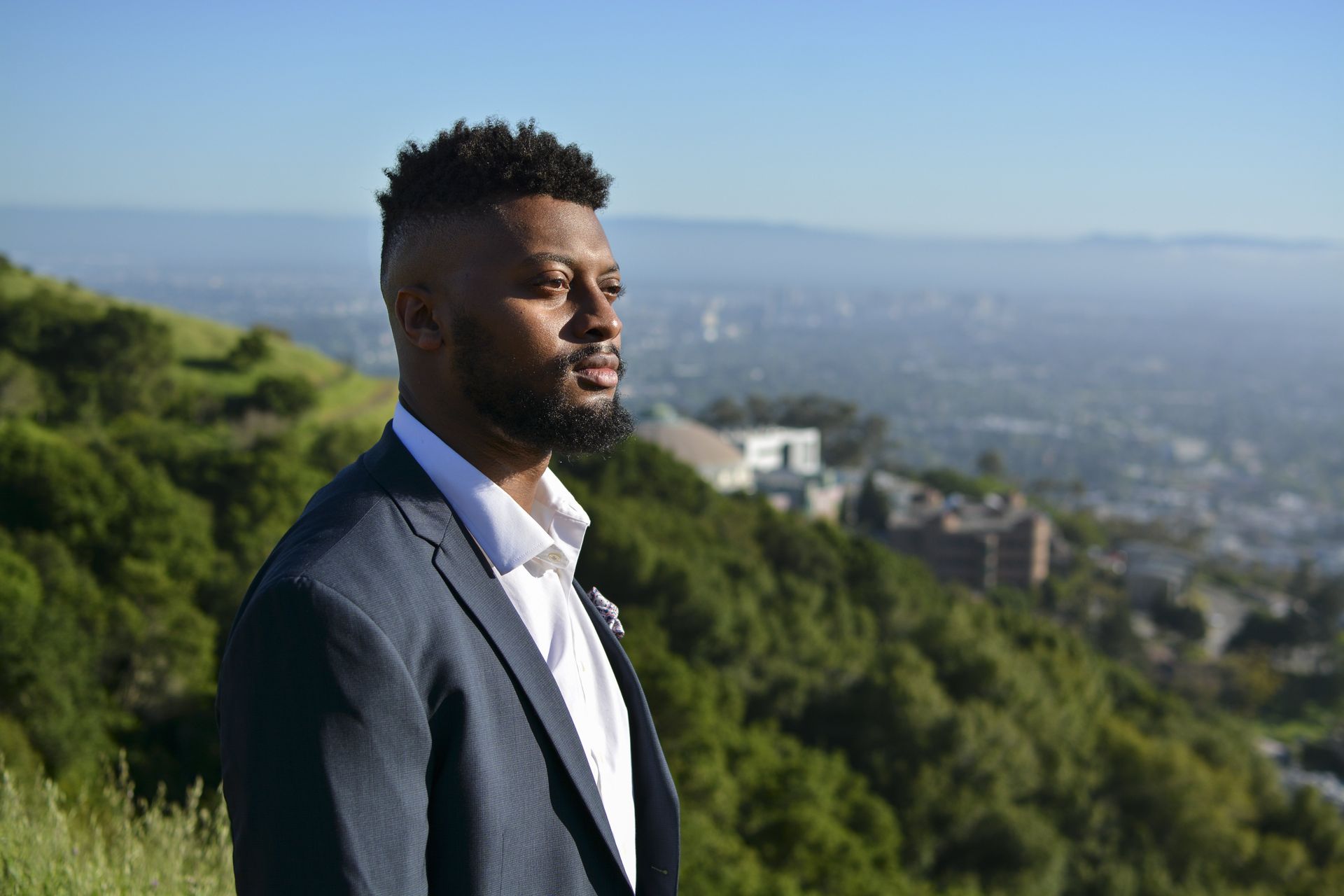 Man in blue suit, looking thoughtfully over a cityscape from a grassy hillside on a sunny day.