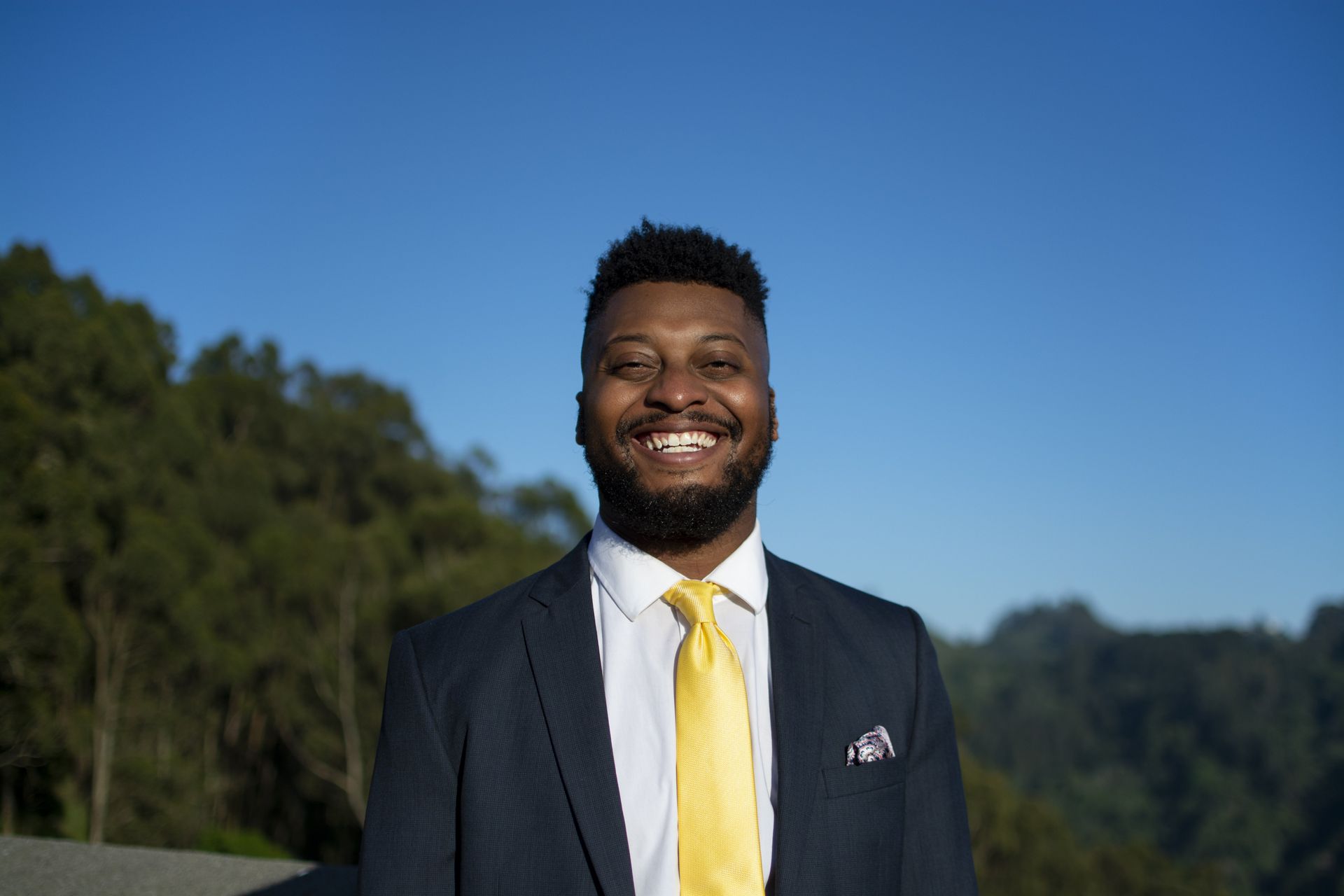Man in suit smiling outdoors with yellow tie, mountains, and blue sky.