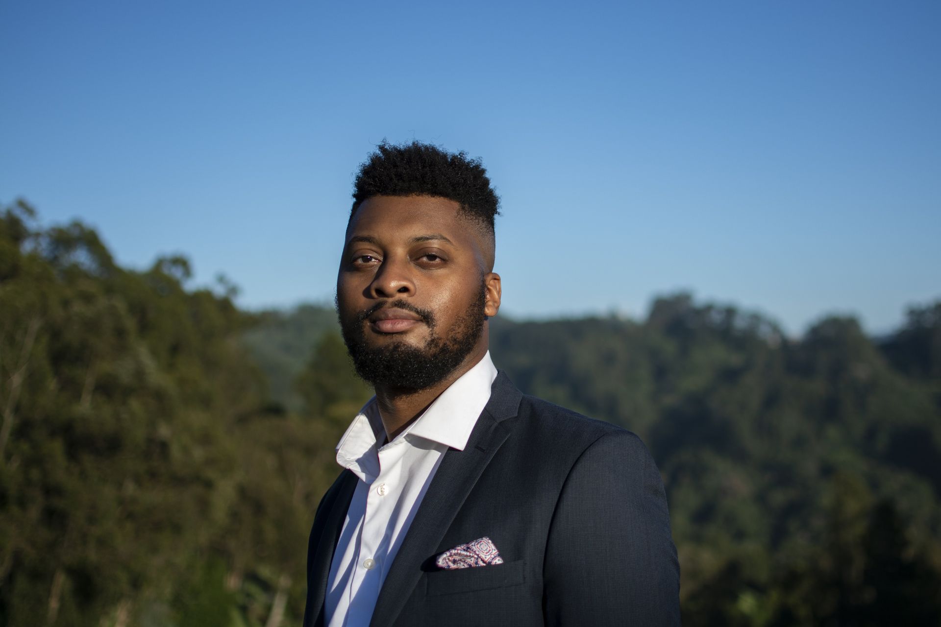 Man in navy blazer and white shirt, smiling, with forest and blue sky background.