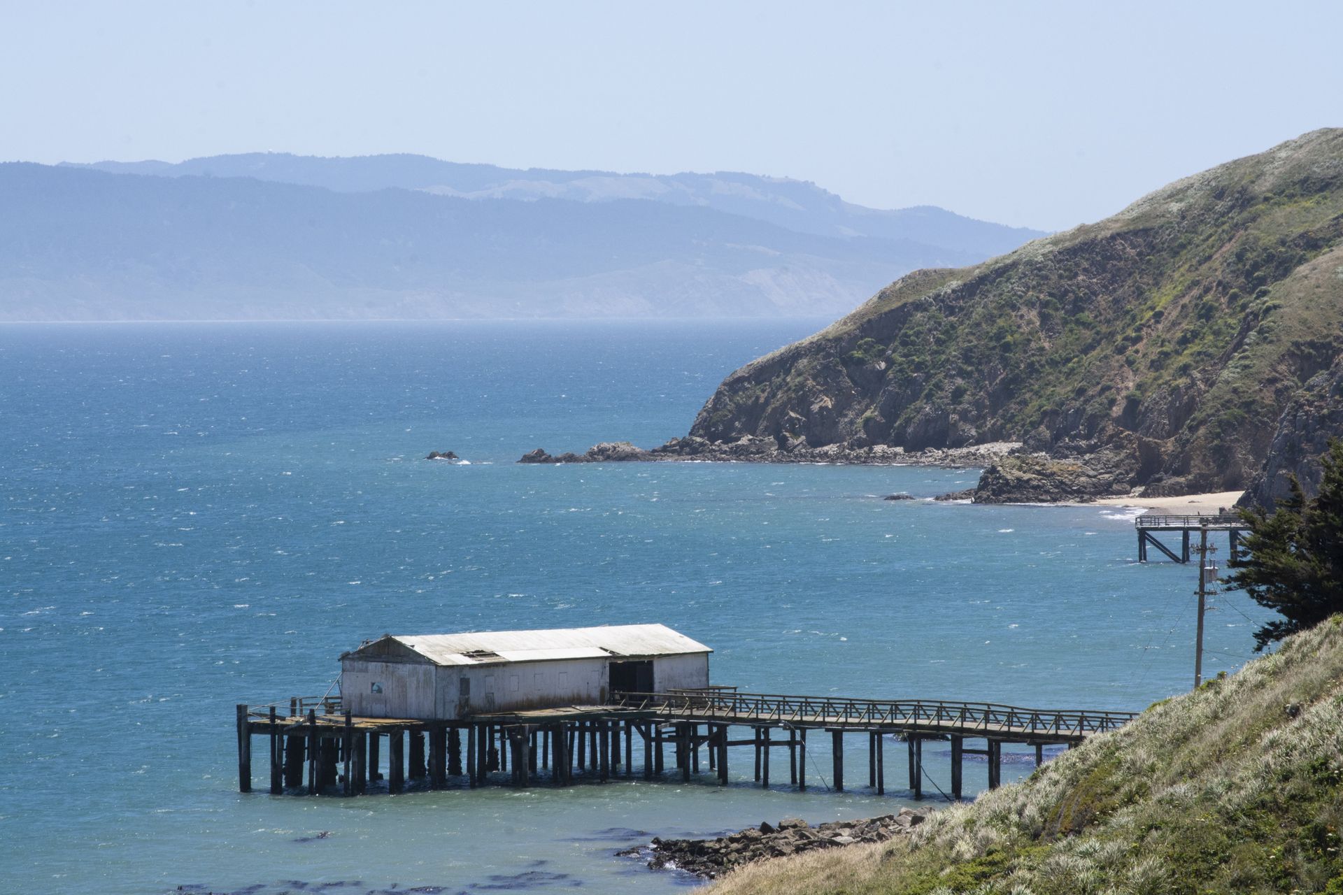 Old wooden pier with a weathered building, blue ocean, and hills in the background.