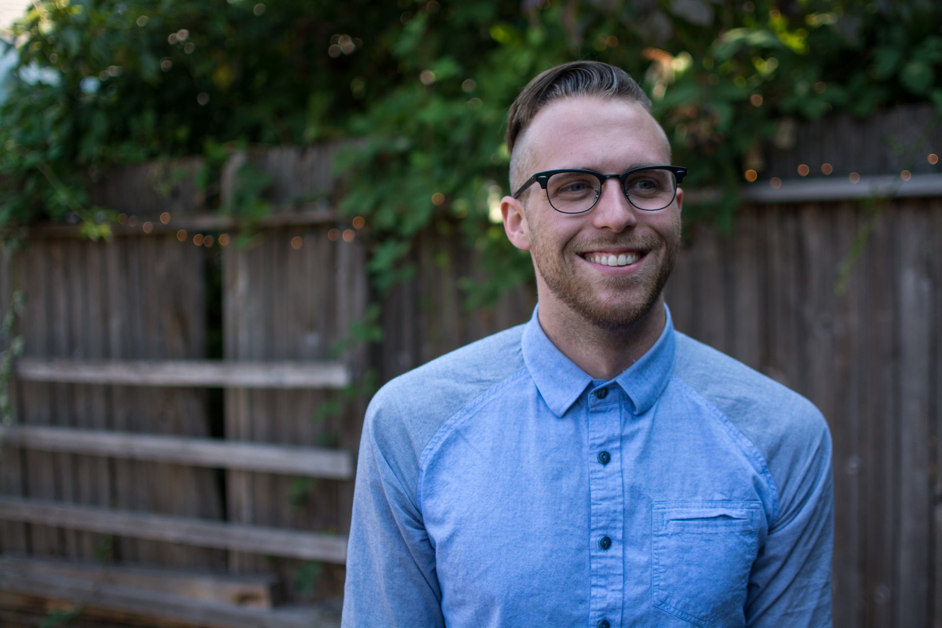 Smiling man with glasses, blue shirt, standing in front of a wooden fence, outdoors.