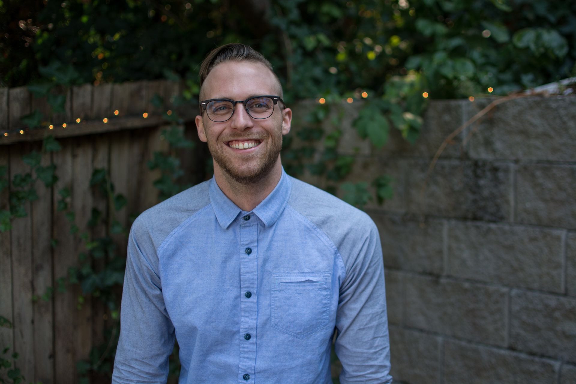 Man with glasses smiles, standing outdoors in front of a fence and stone wall. He wears a blue shirt.