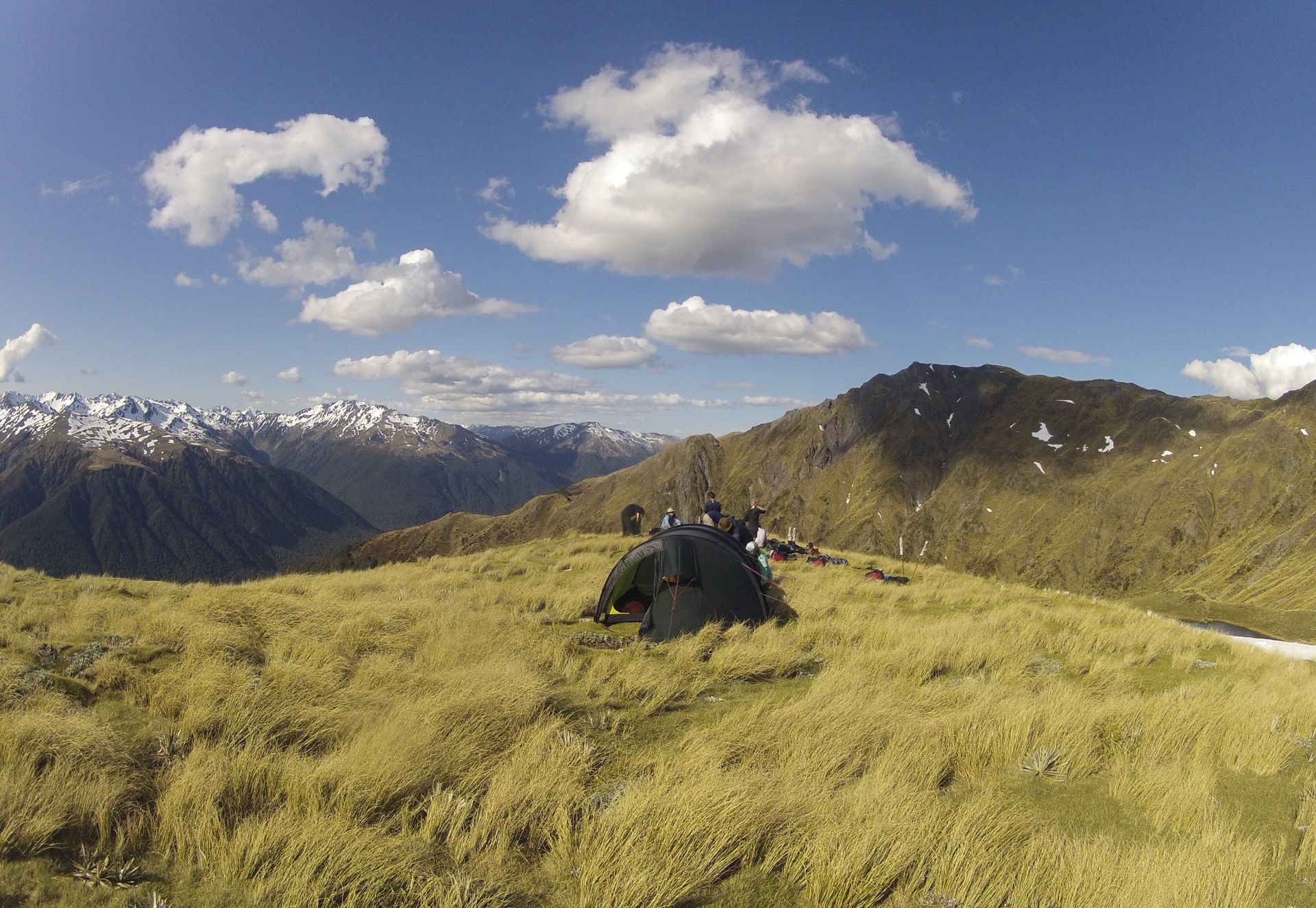 Green grassy mountain ridge with people; snowy peaks in the background, blue sky with white clouds.