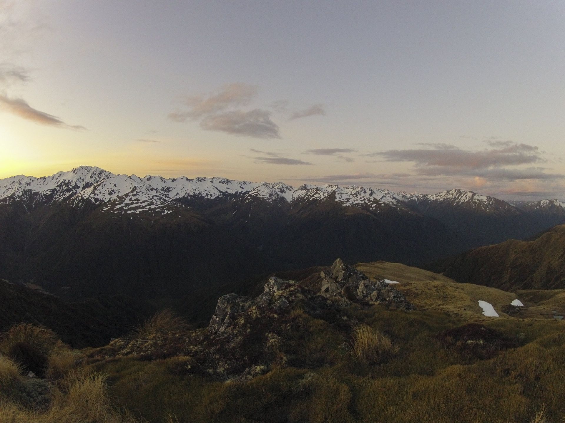 Mountain range at dusk, with snow-covered peaks under a dusky sky, overlooking a valley.