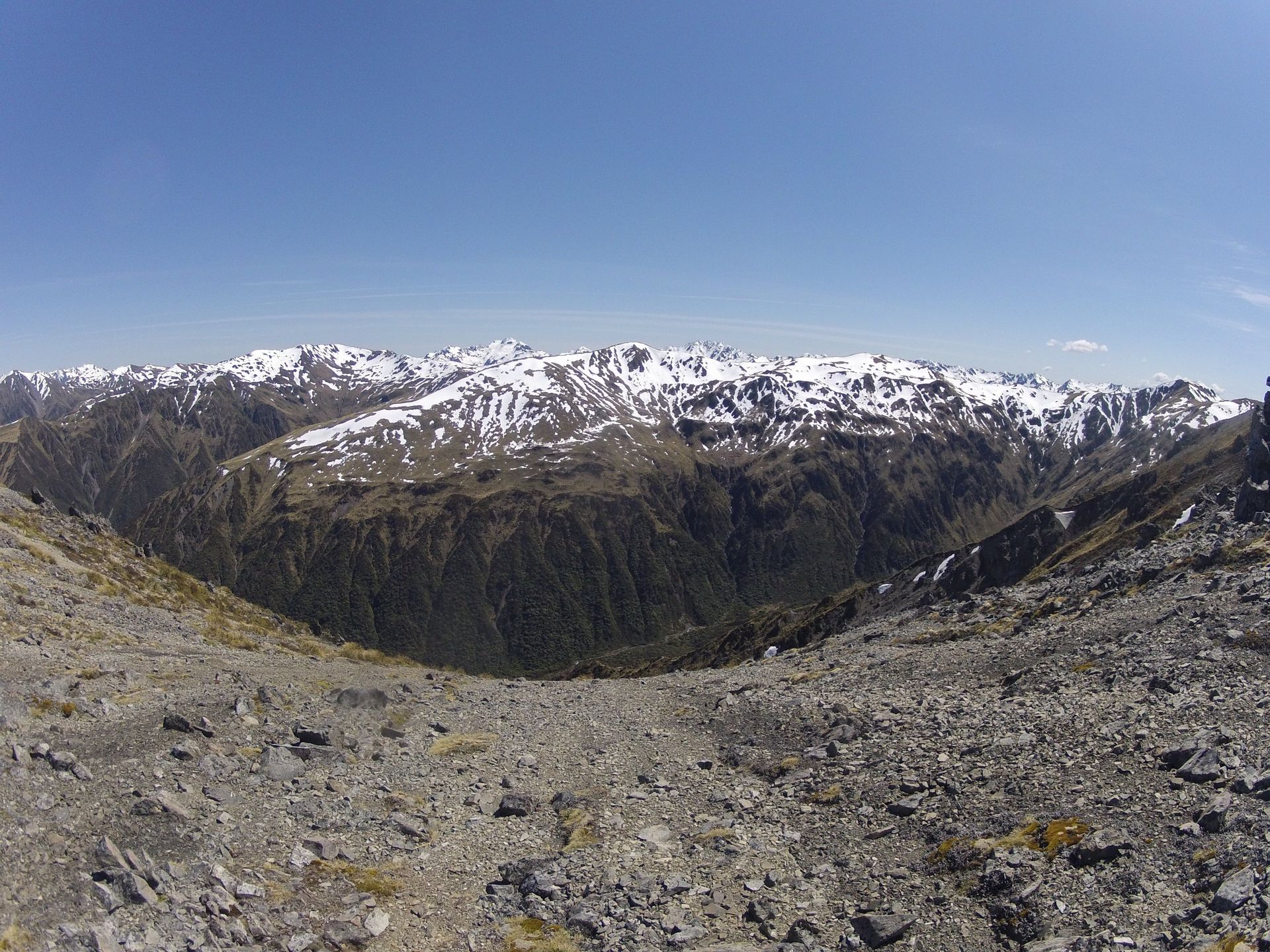 Rocky terrain slopes down to a valley with snow-capped mountains under a clear blue sky.