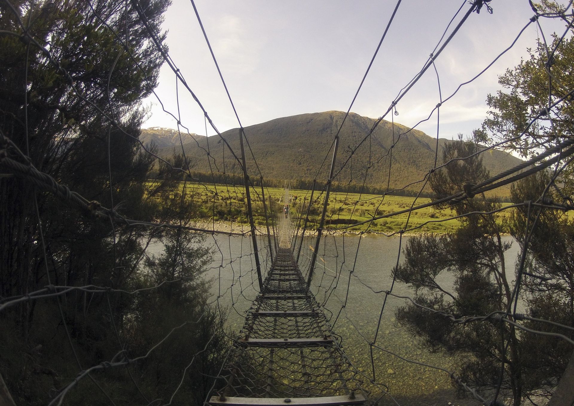 Suspension bridge over water, view toward a hillside under a sunny sky.
