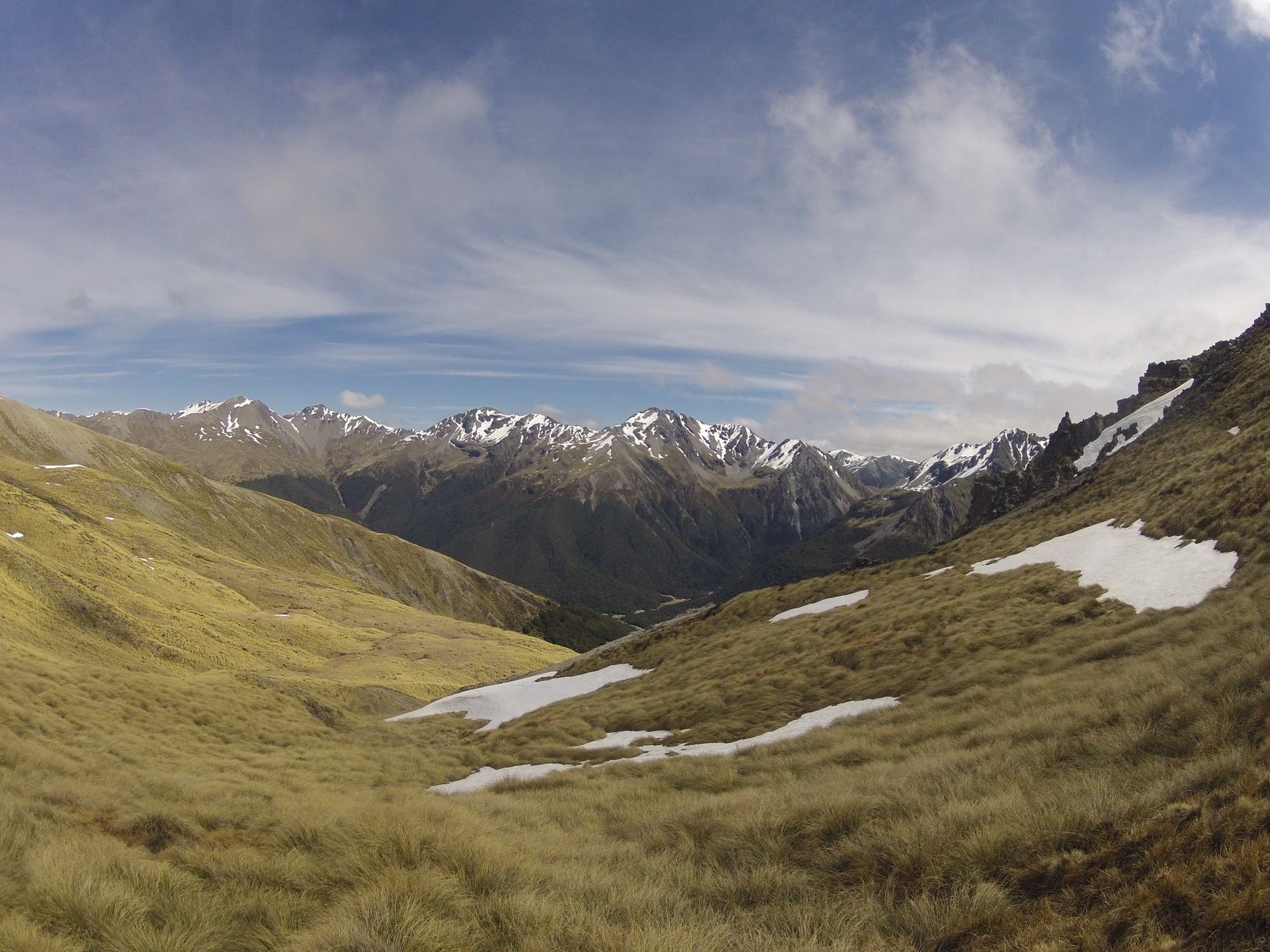 Valley with grassy slopes and patches of snow leading to snow-capped mountains under a blue sky.
