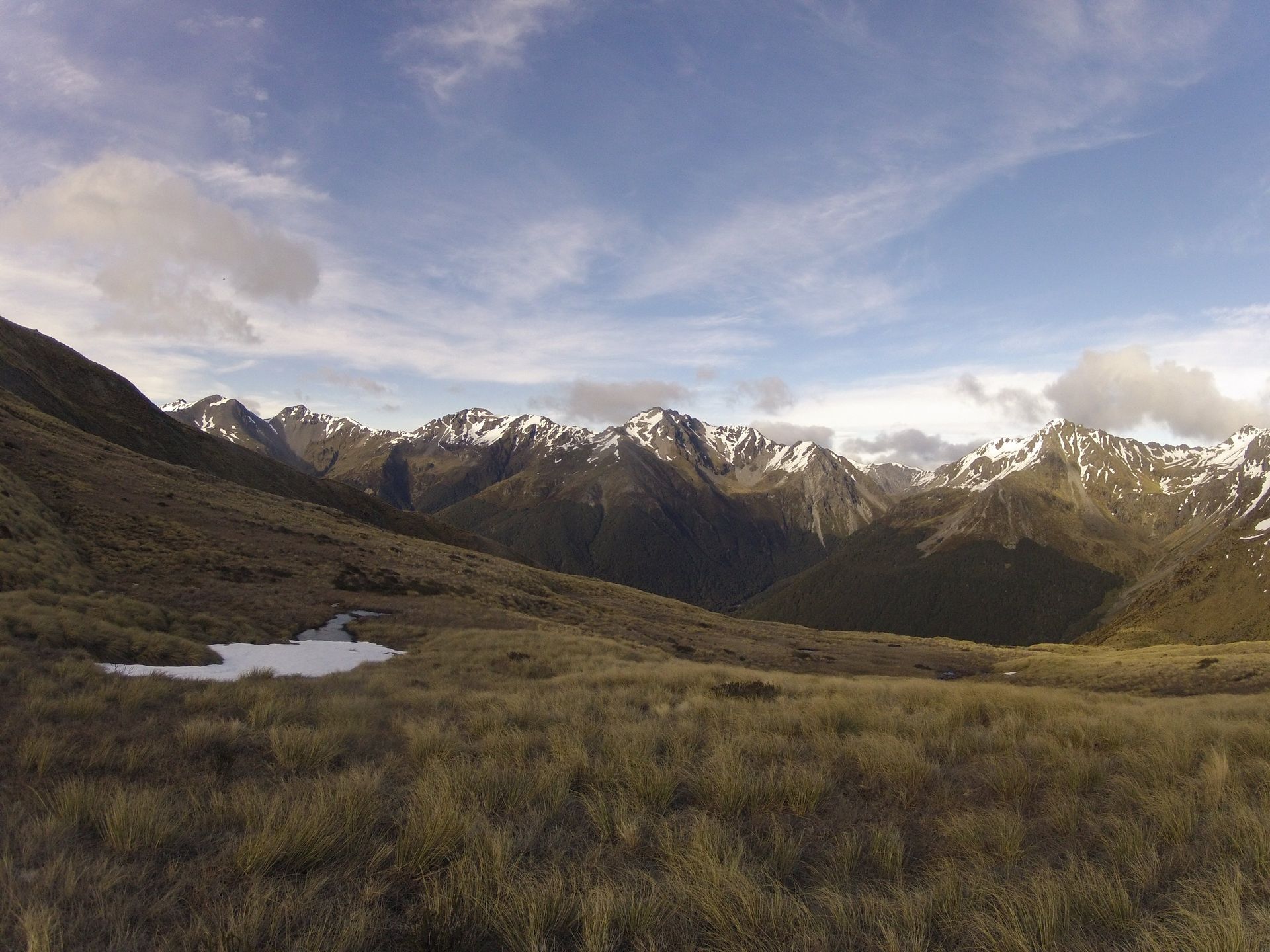 Grassy valley with mountains in the background under a partly cloudy sky. Some snow on the mountain peaks.