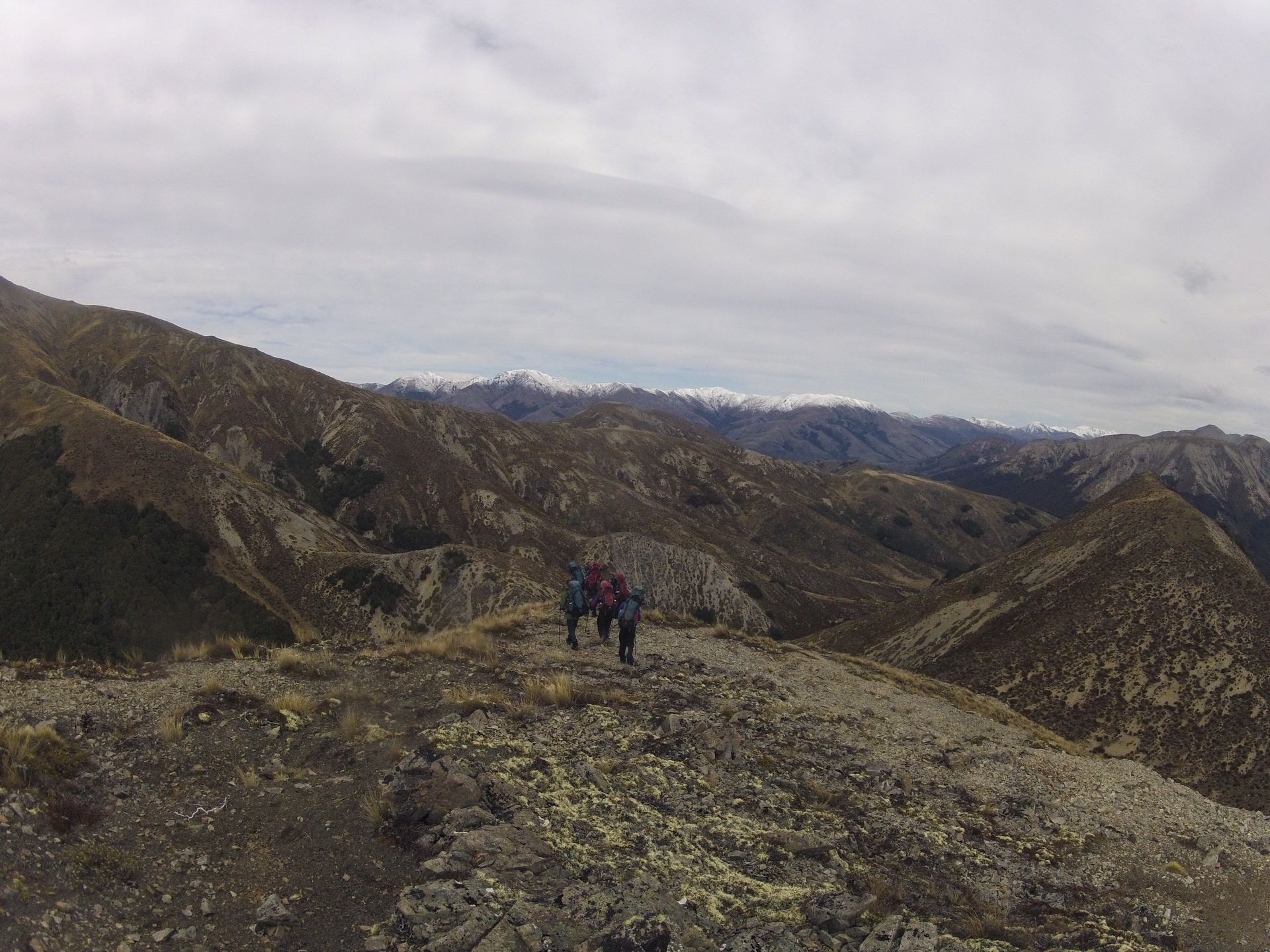 Hikers on a rocky mountain ridge with distant snow-capped peaks under a cloudy sky.