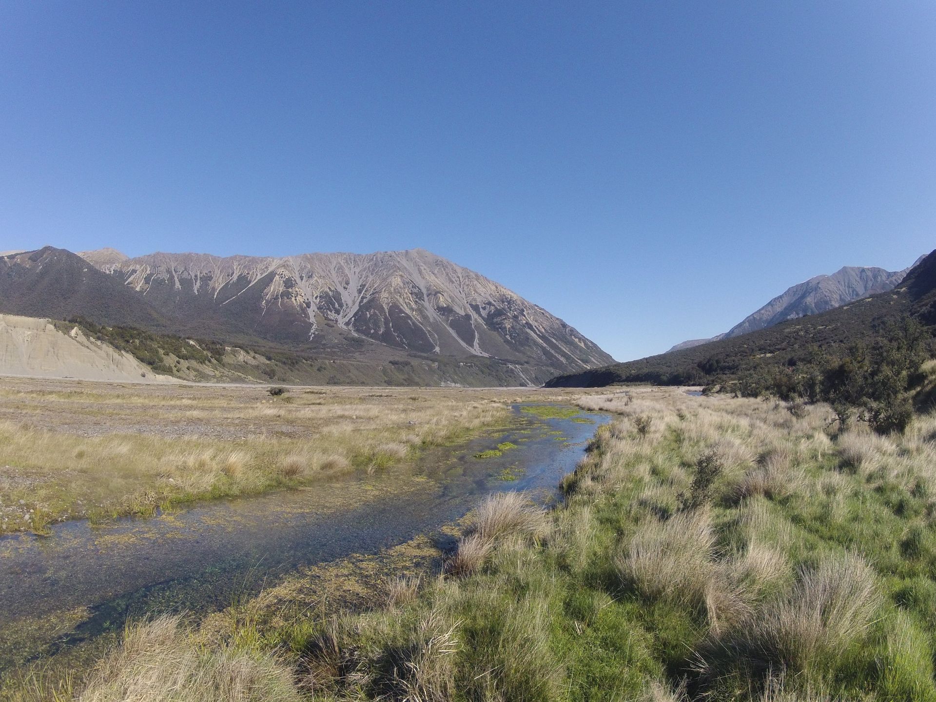 A stream meanders through a grassy plain toward mountains under a clear blue sky.