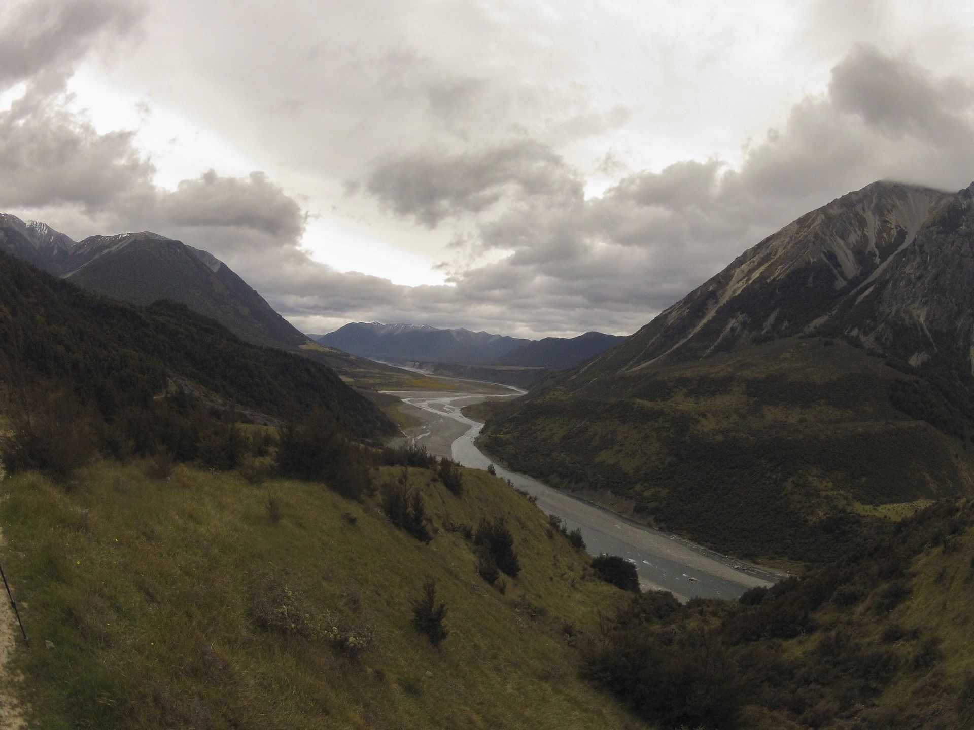 Valley with river, mountains, and overcast sky. Green hills and forest frame the scene.