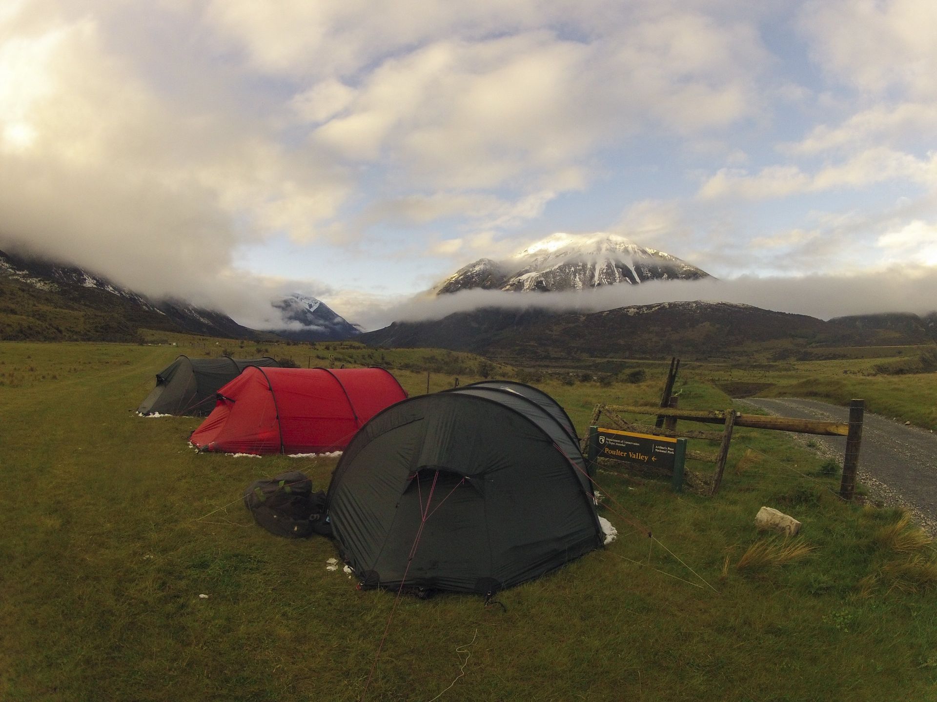 Three tents on a green field with mountains in the background, partially covered in snow and clouds.