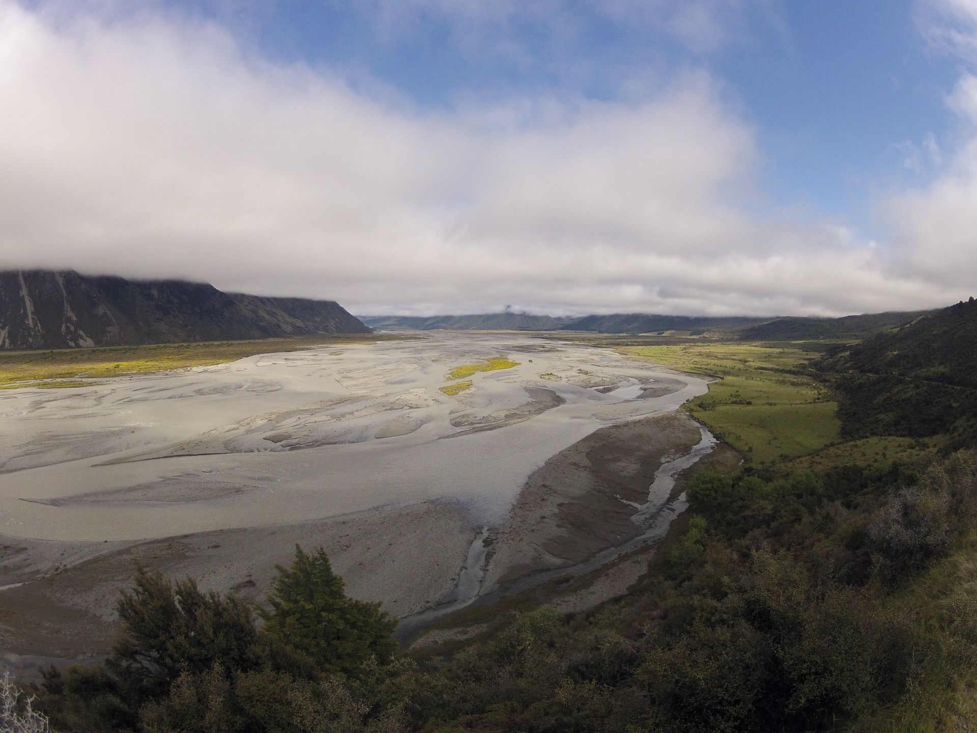 Wide valley with a pale, braided river flowing through; green vegetation on the edges, under a cloudy sky.