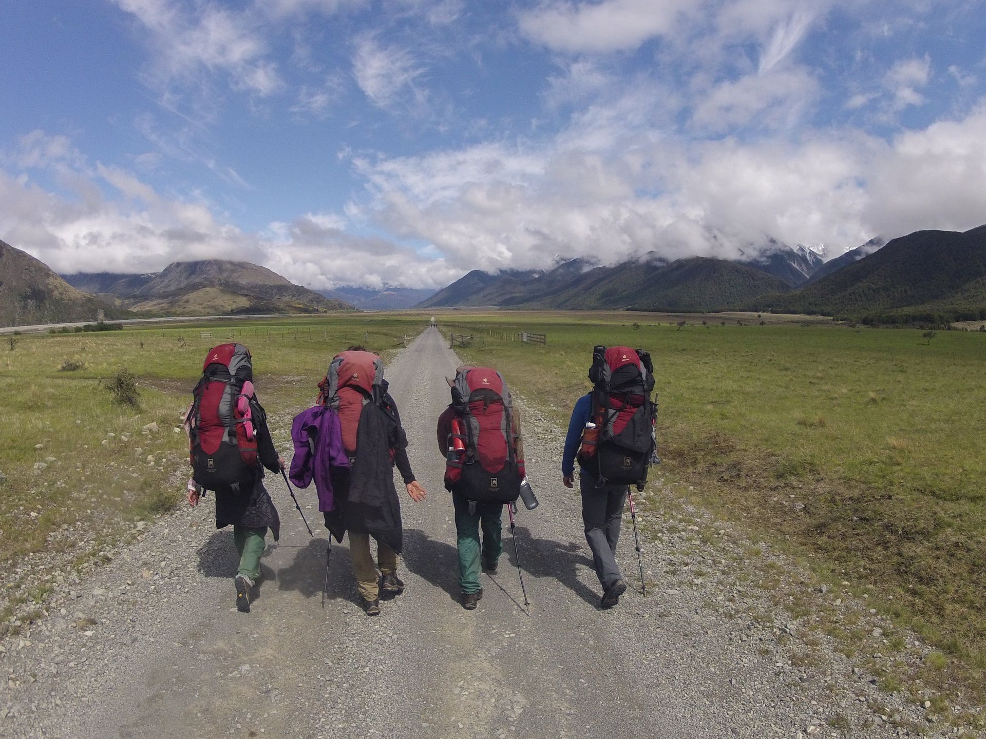 Four hikers with large backpacks walk down a gravel path towards mountains on a sunny day.