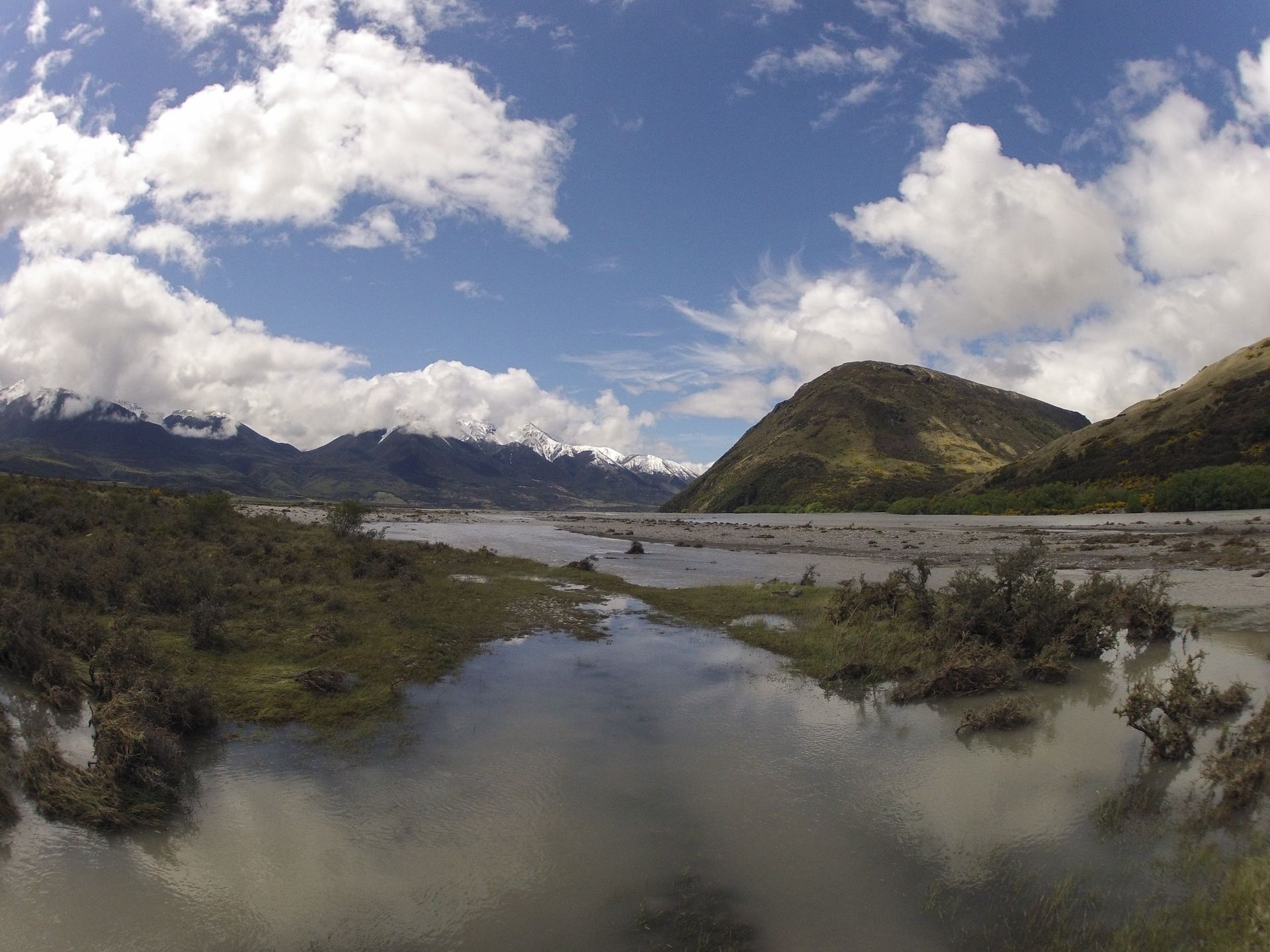 Wide shot of a river flowing through a valley, mountains in the background, blue sky with clouds.