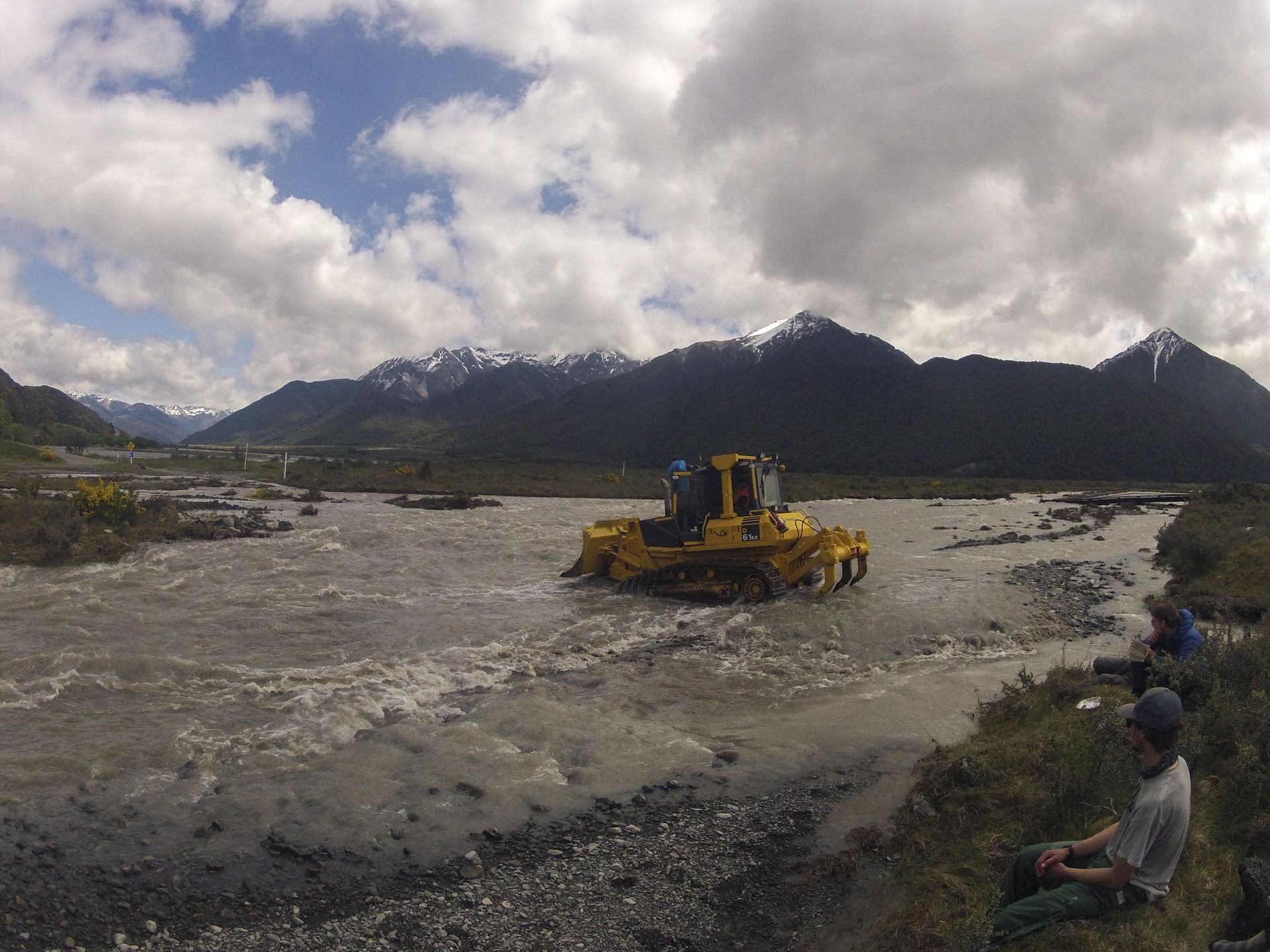 Yellow tracked vehicle fording a river, mountains in the background, cloudy sky. Two people watch from the shore.