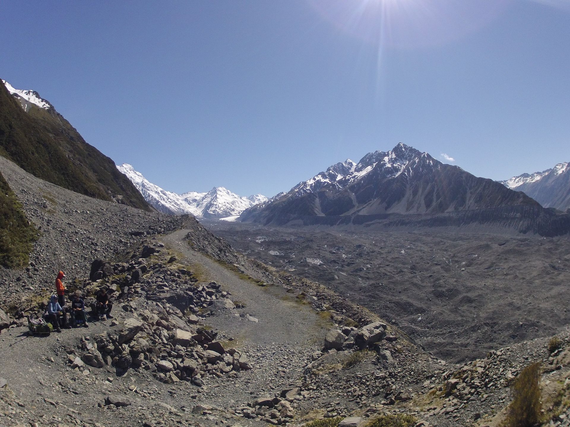Hikers on a rocky path with a glacier and snow-capped mountains under a bright blue sky.