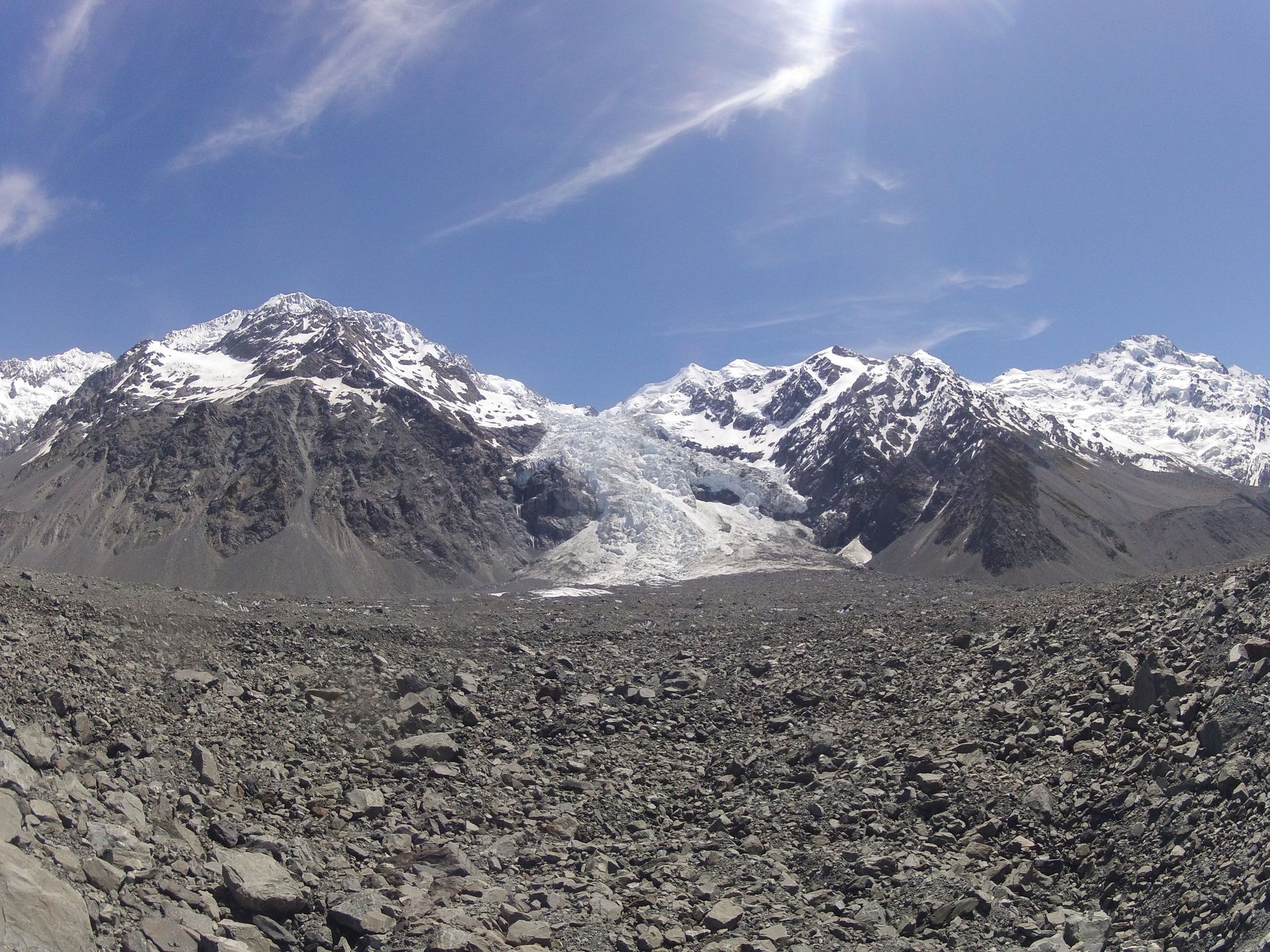 Mountains with snow-capped peaks and a glacier, seen from a rocky, gray terrain under a blue sky.