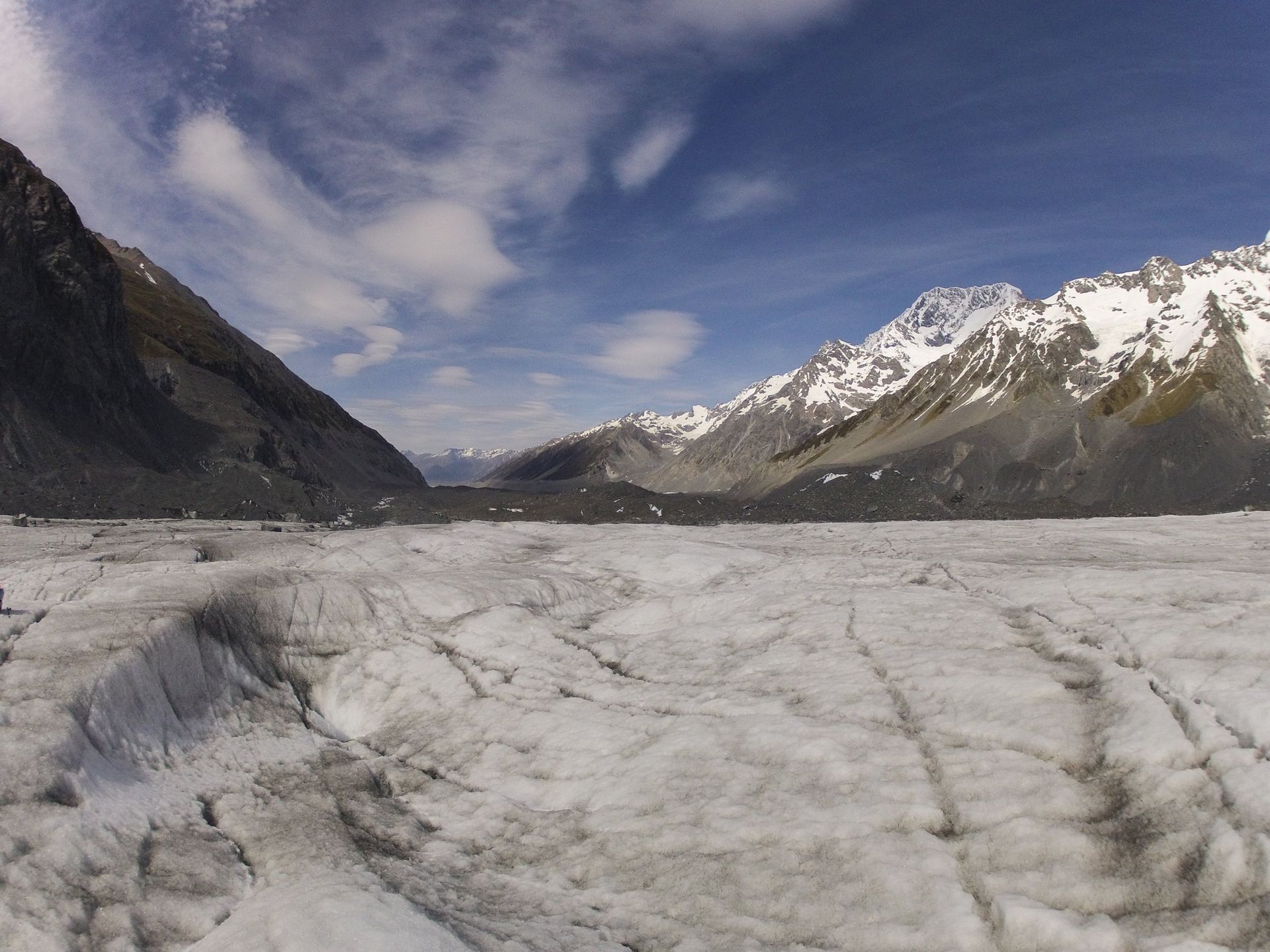 Glacial landscape: ice and crevasses in valley between snow-capped mountains under partly cloudy blue sky.