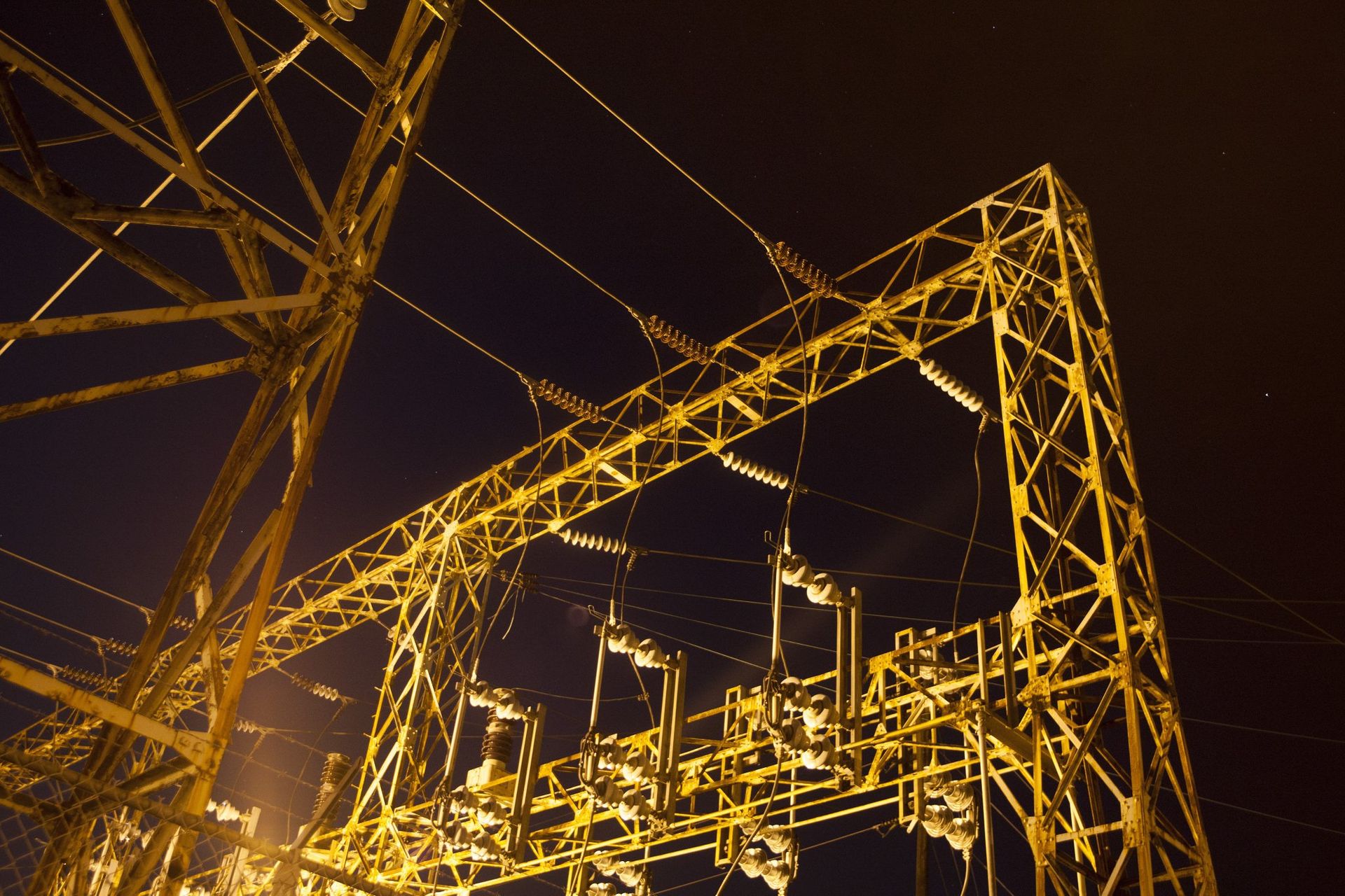 Night view of a lit electrical substation, with metal framework and power lines against a dark sky.