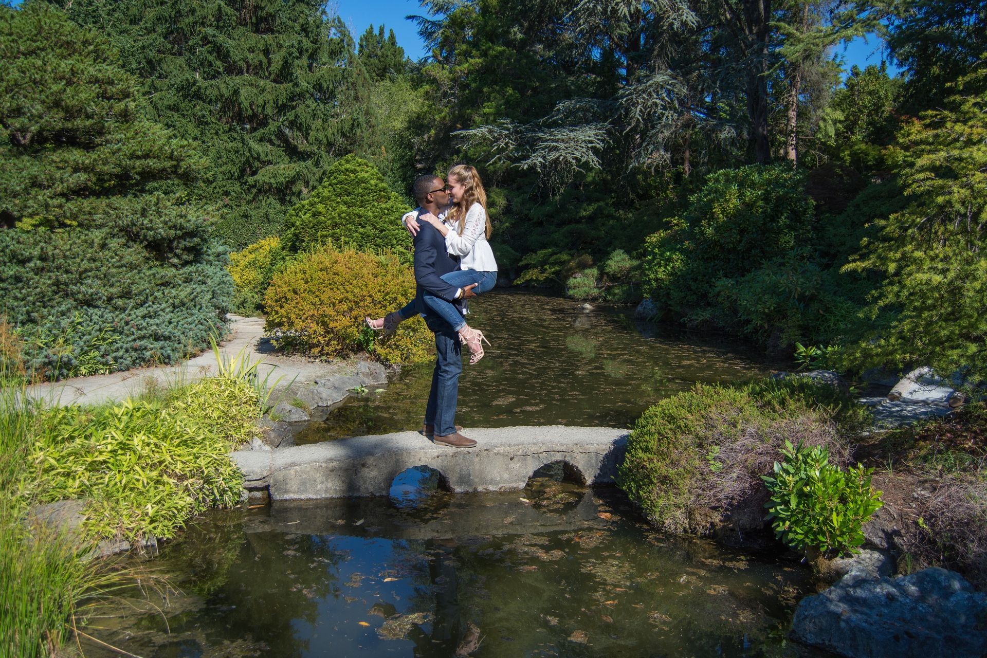 Couple embracing on a small stone bridge over a pond, surrounded by greenery.
