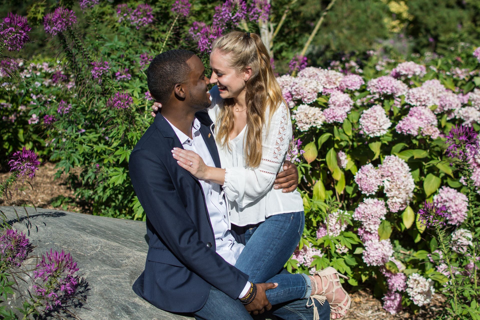 Couple embracing among pink flowers; smiling faces, he wears a blazer, she wears a white top.