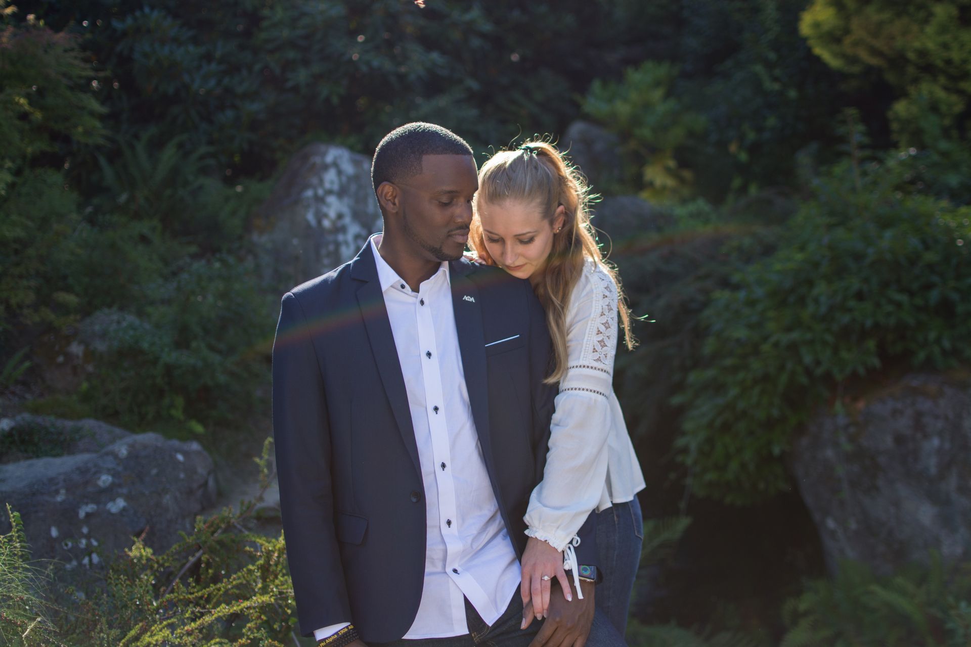 Couple embracing outdoors; woman's head on man's shoulder. Lush greenery surrounds them.