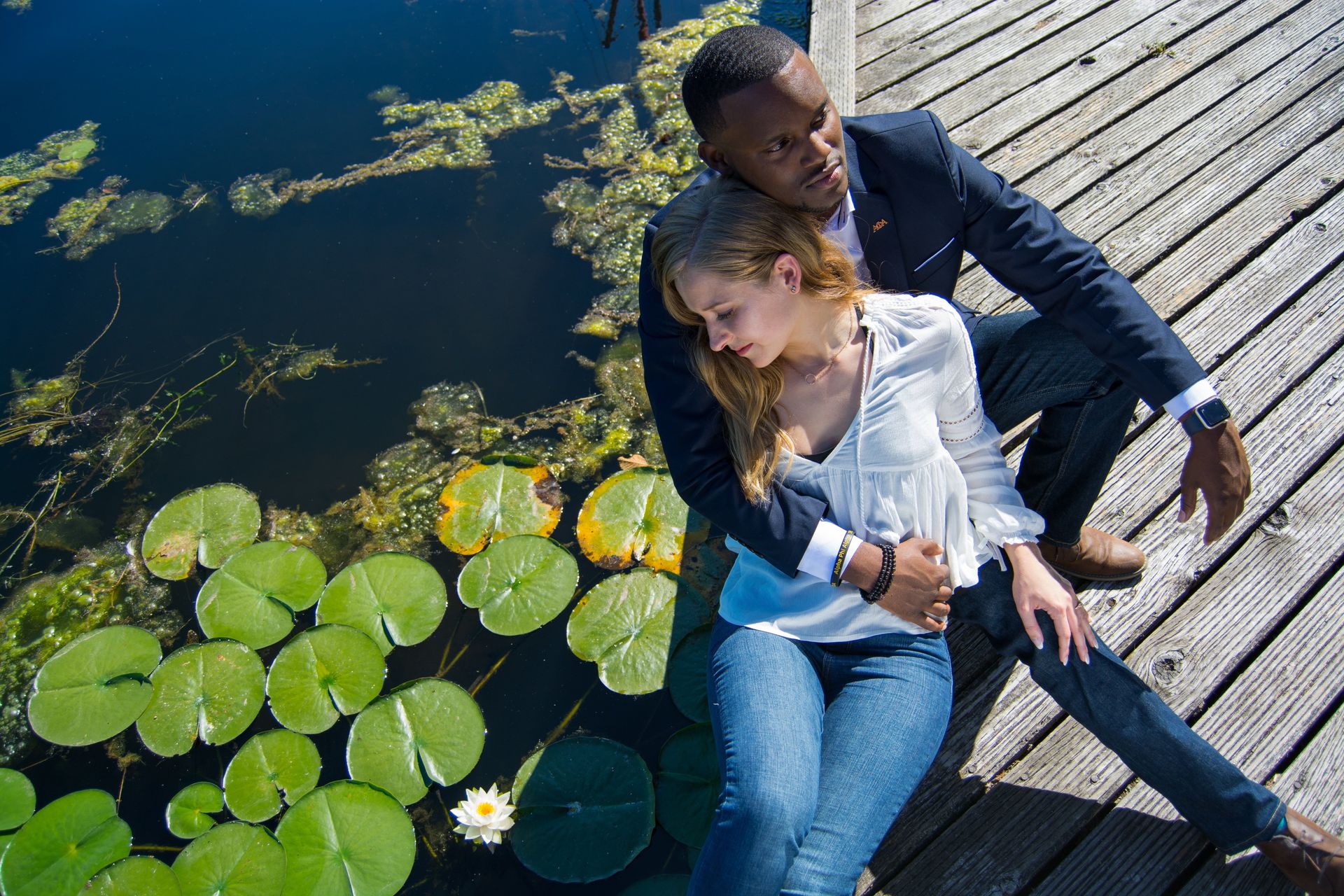 Man in blazer embraces woman on wooden dock next to lily pads.
