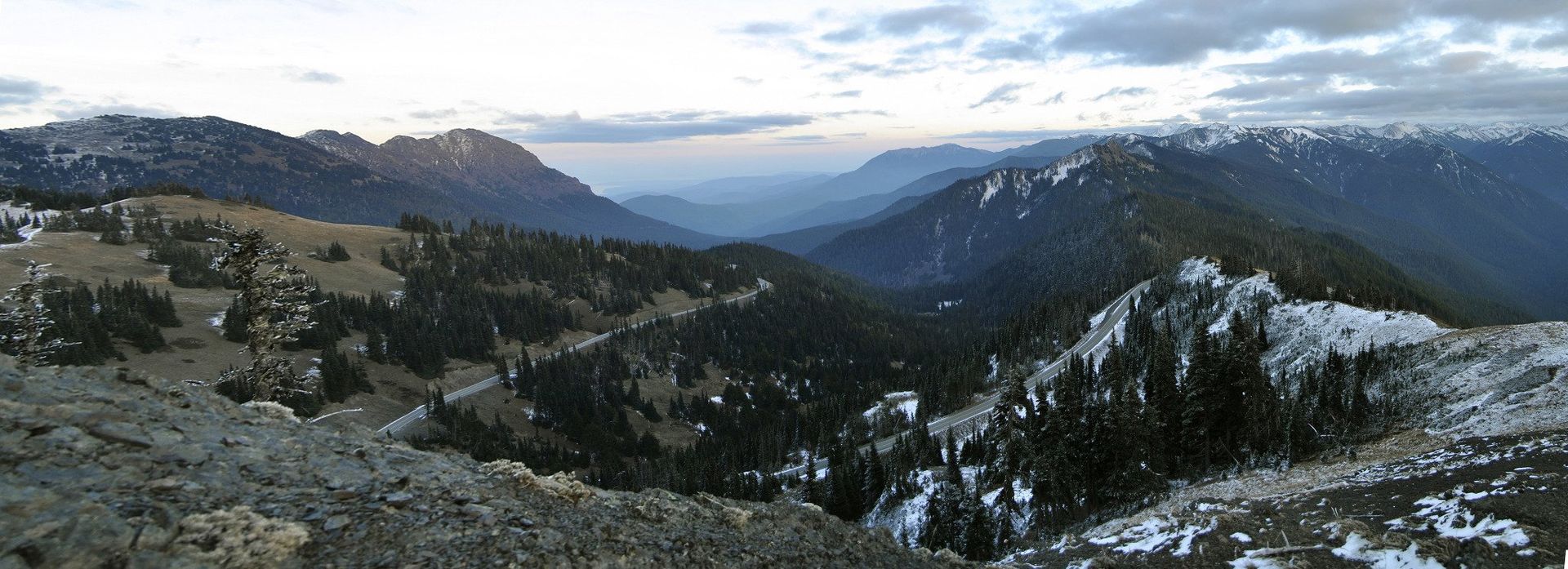 Panoramic view of a mountain valley with snow-capped peaks and evergreen trees under a cloudy sky.