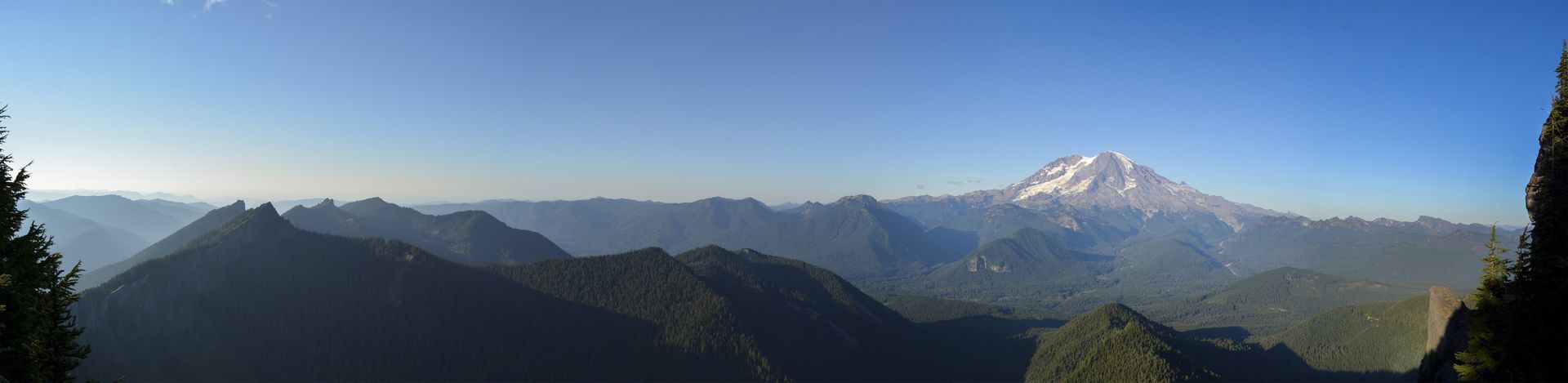 Panoramic view of forested mountains with a snow-capped peak under a clear blue sky.