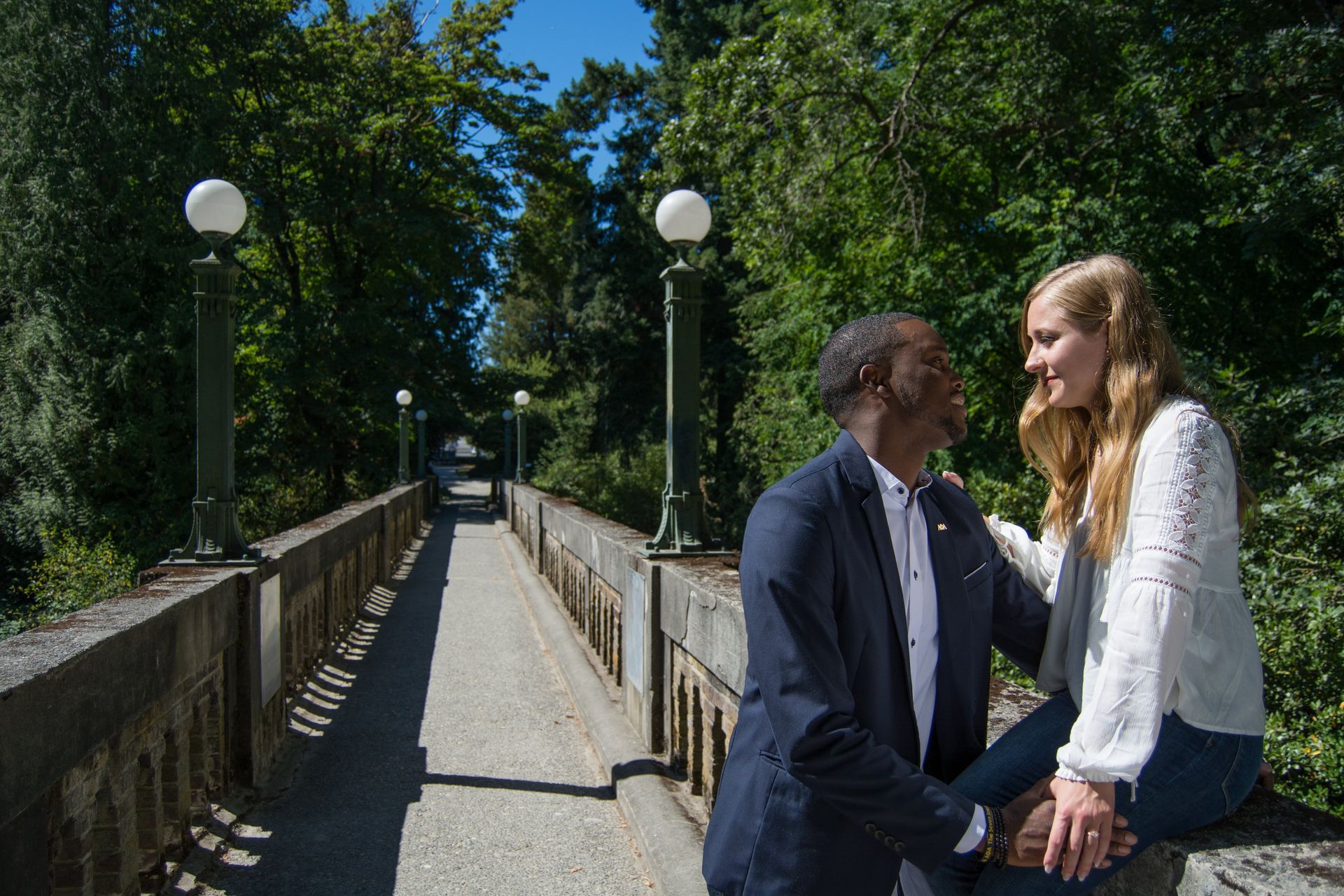 Man and woman on a bridge, smiling, trees in background, sunny day.