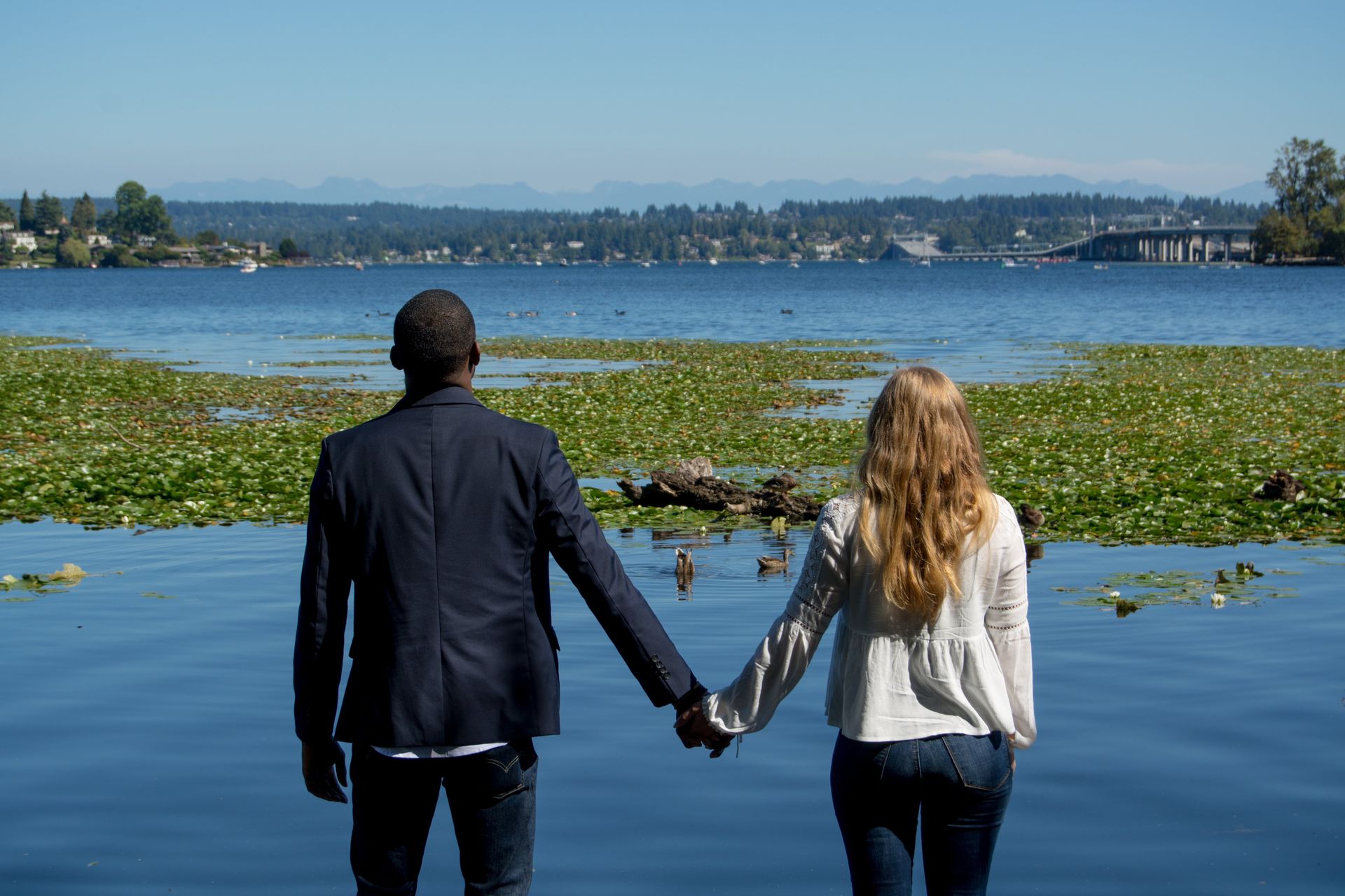 Couple holding hands, facing lake. Man in dark jacket, woman with blonde hair, sunny day.