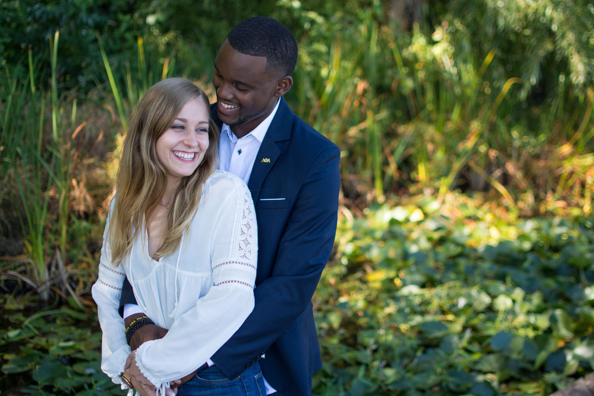 Couple embraces outdoors, woman smiles, man in suit hugs her, greenery background.