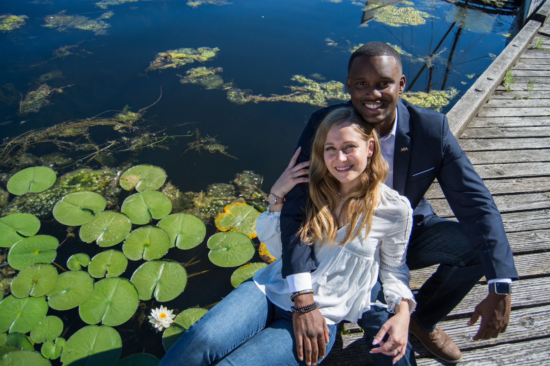 Couple sitting on a wooden dock by a pond with lily pads. Man has arm around woman. Sunny day.