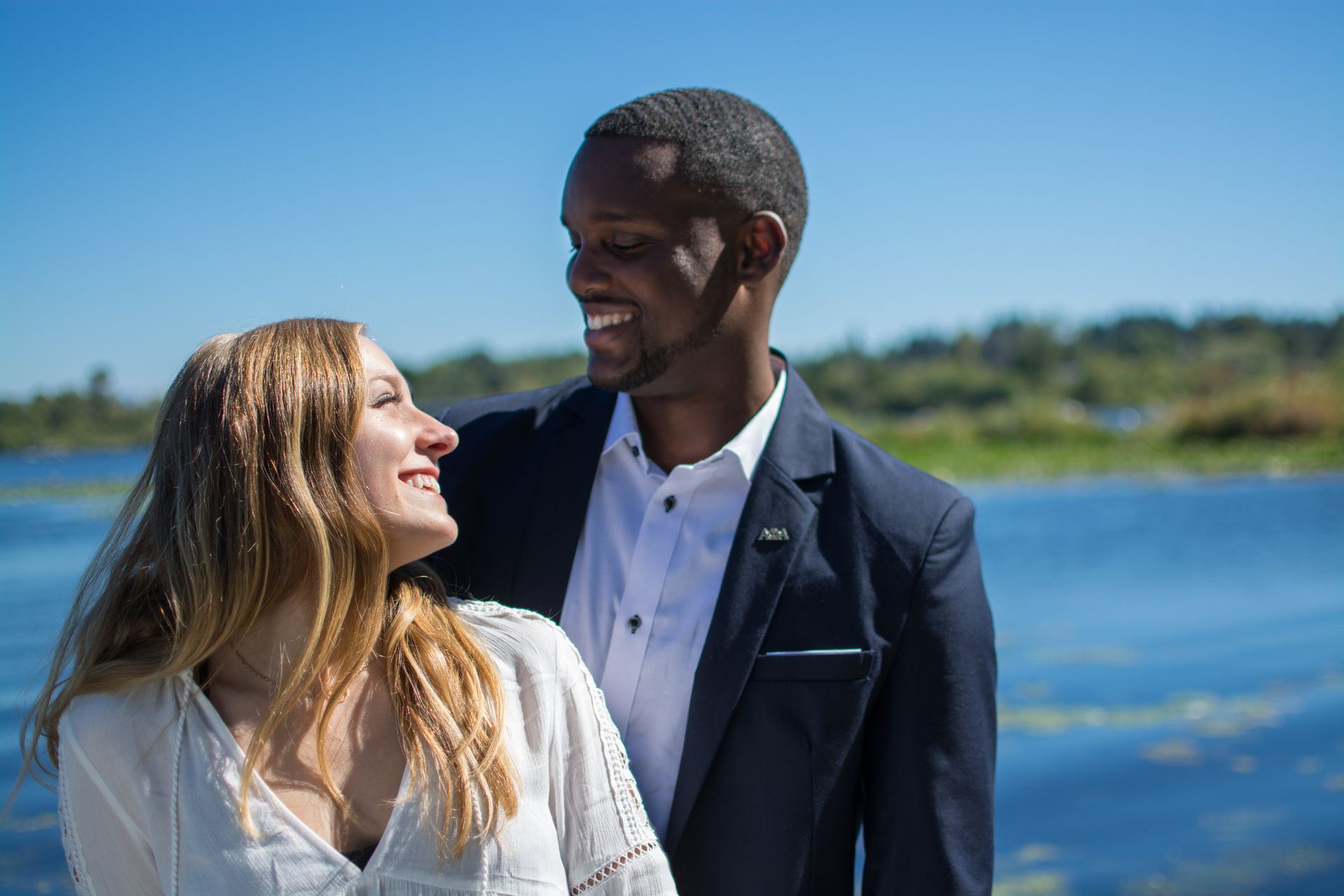 Couple smiling, looking at each other, blue water backdrop. Man in navy blazer, woman in white blouse.