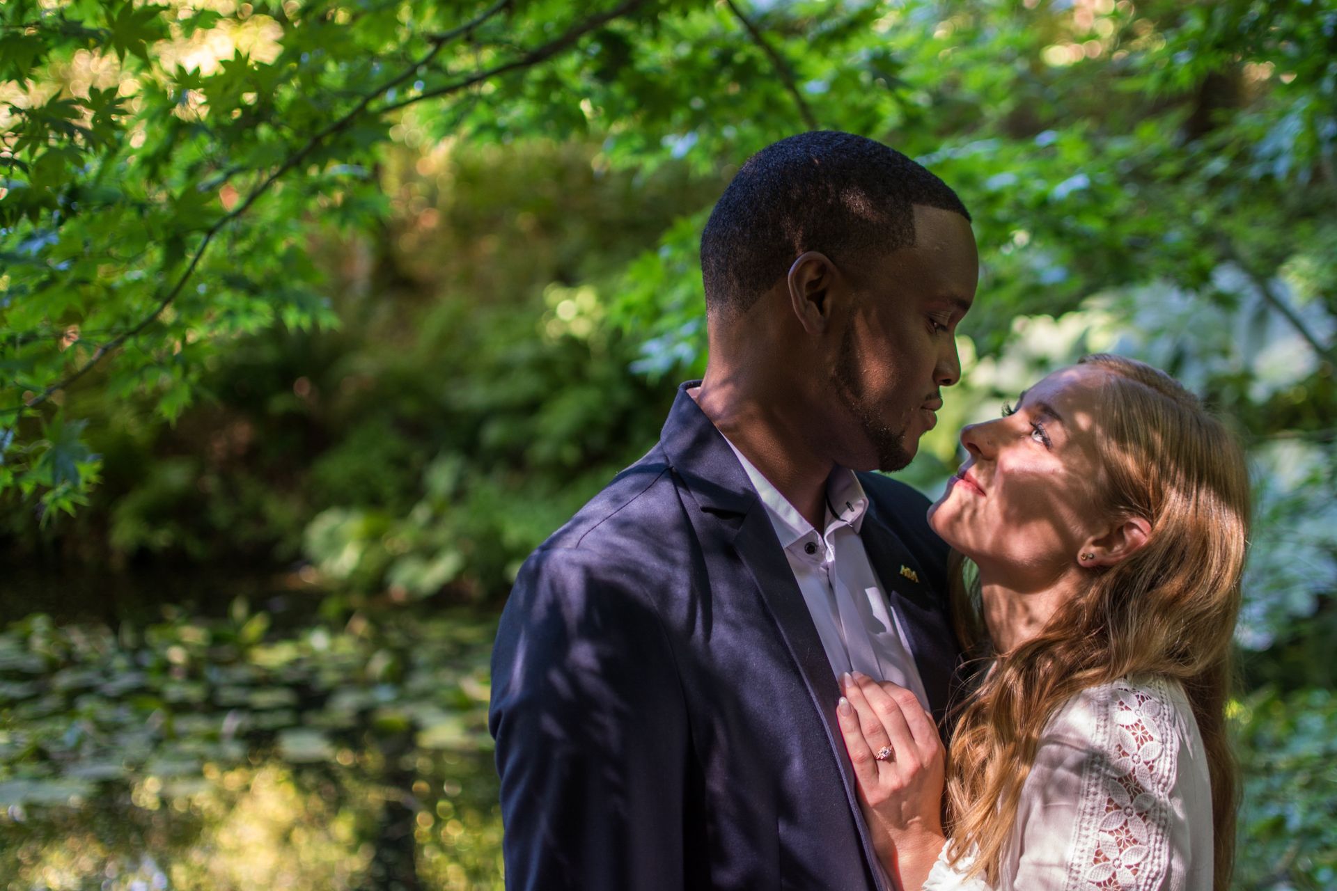 Couple embracing, in a garden. Man in a suit jacket, woman in white, looking at each other near water.