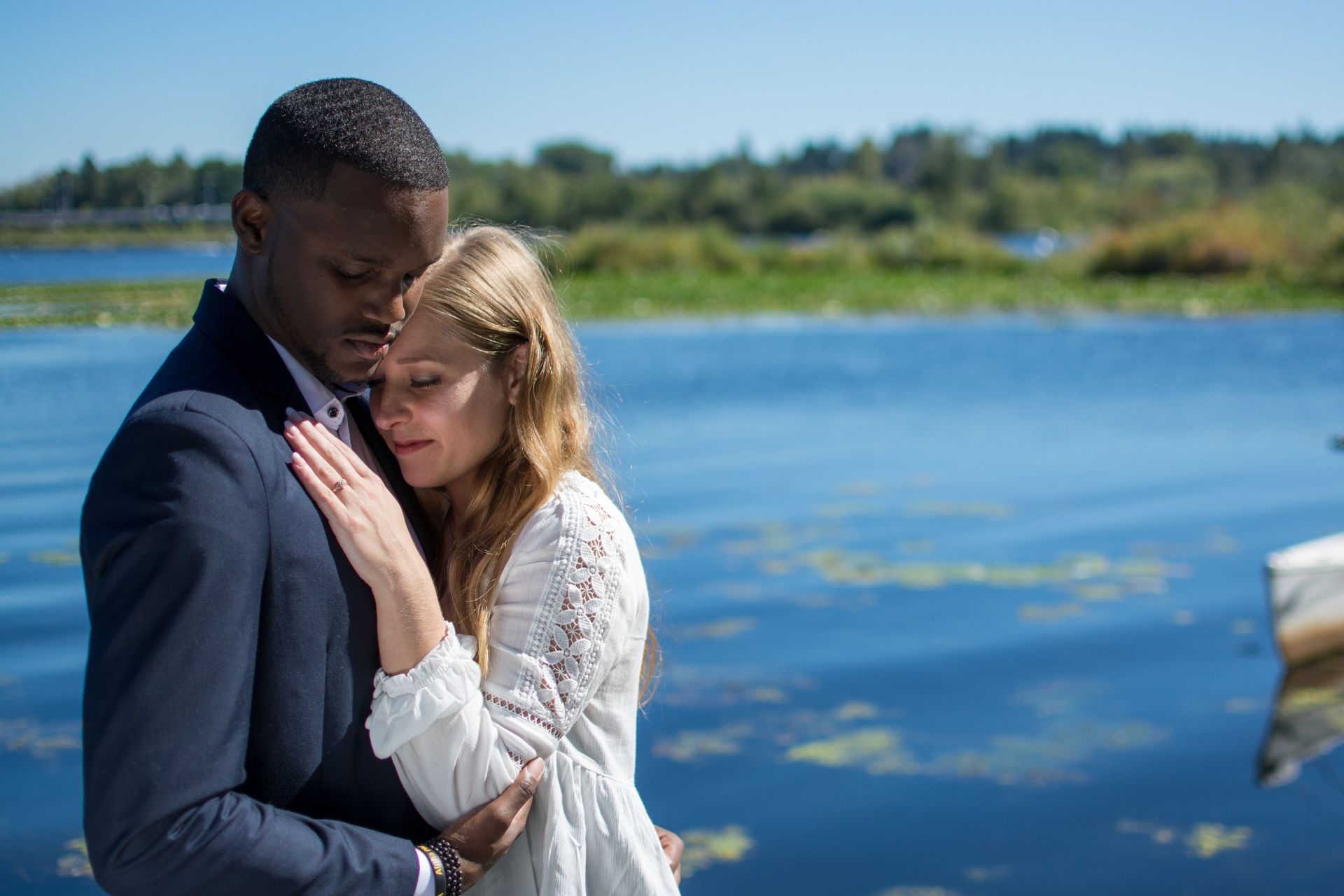 Man in blue suit embraces woman in white dress near a lake.