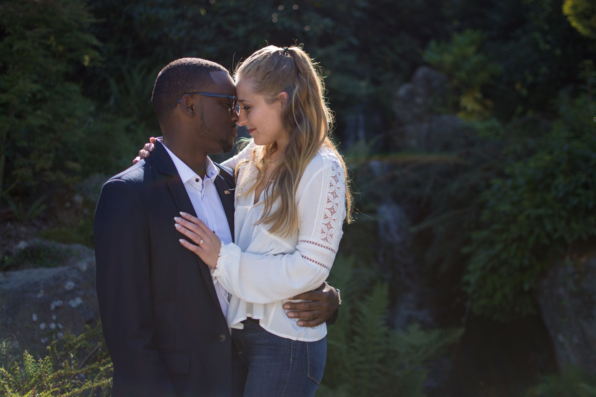 Couple embracing, nose-to-nose, outdoors near waterfall. Man in suit, woman in blouse, both smiling.