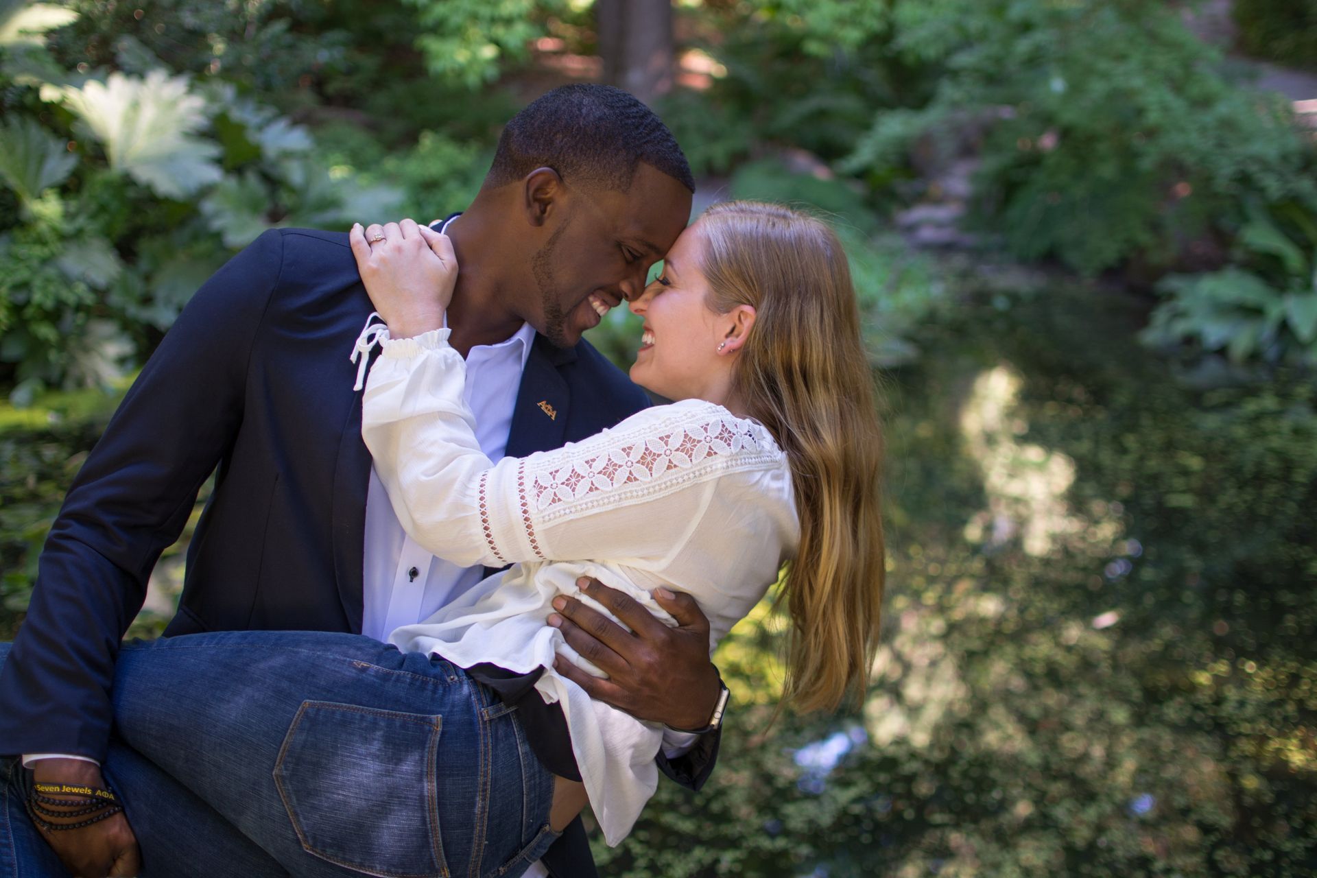 Man holding woman, smiling, in front of pond, outdoors.