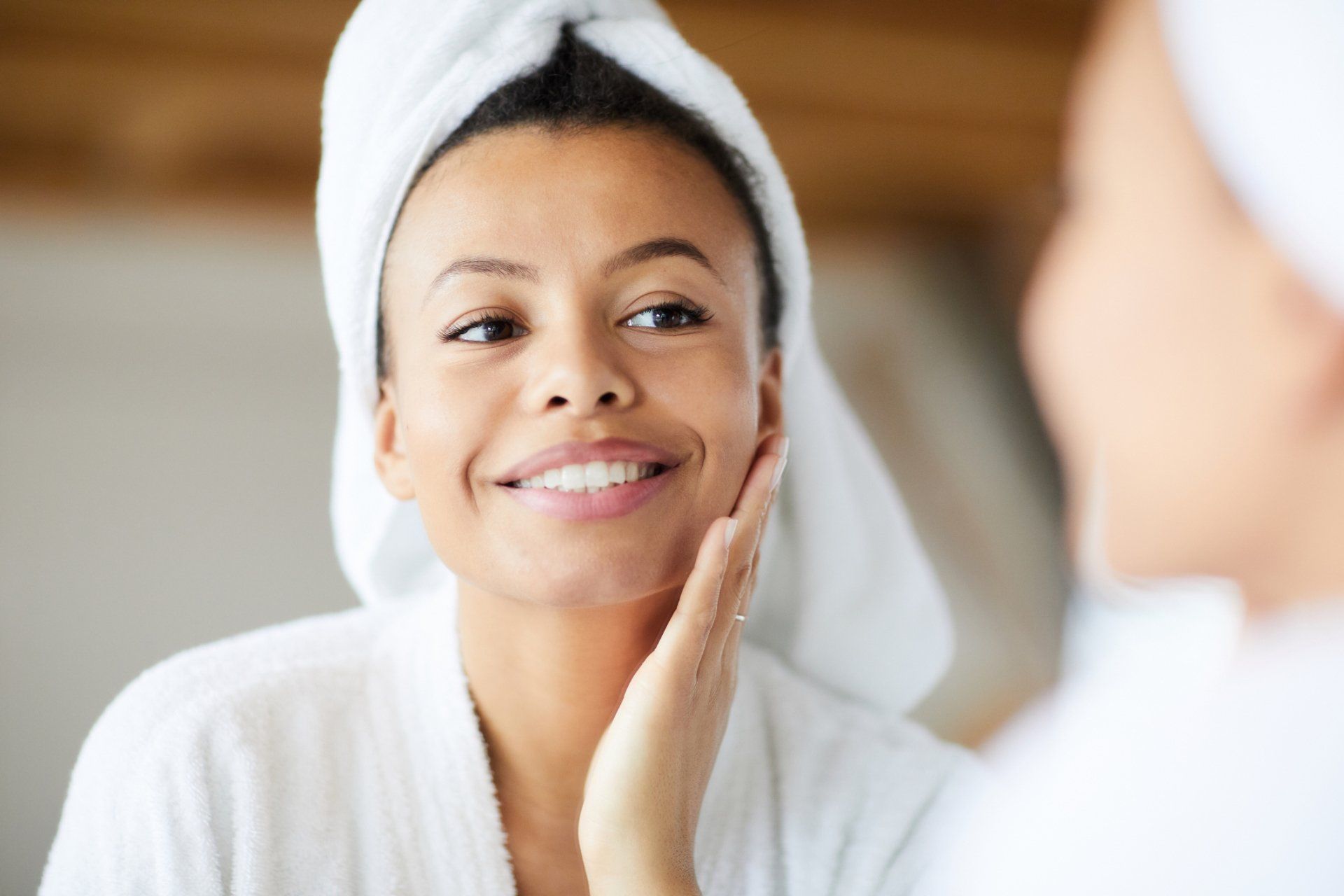 Woman in a white robe and towel looks in a mirror, touching her cheek, smiling.