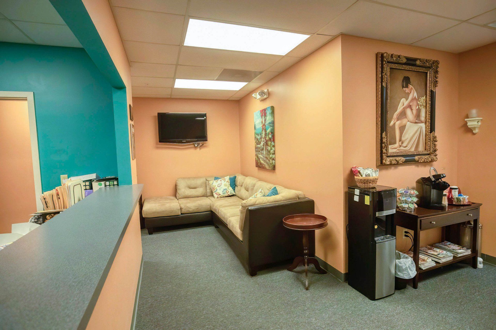 Reception area with teal and peach walls, gray sofa, coffee station, and artwork.