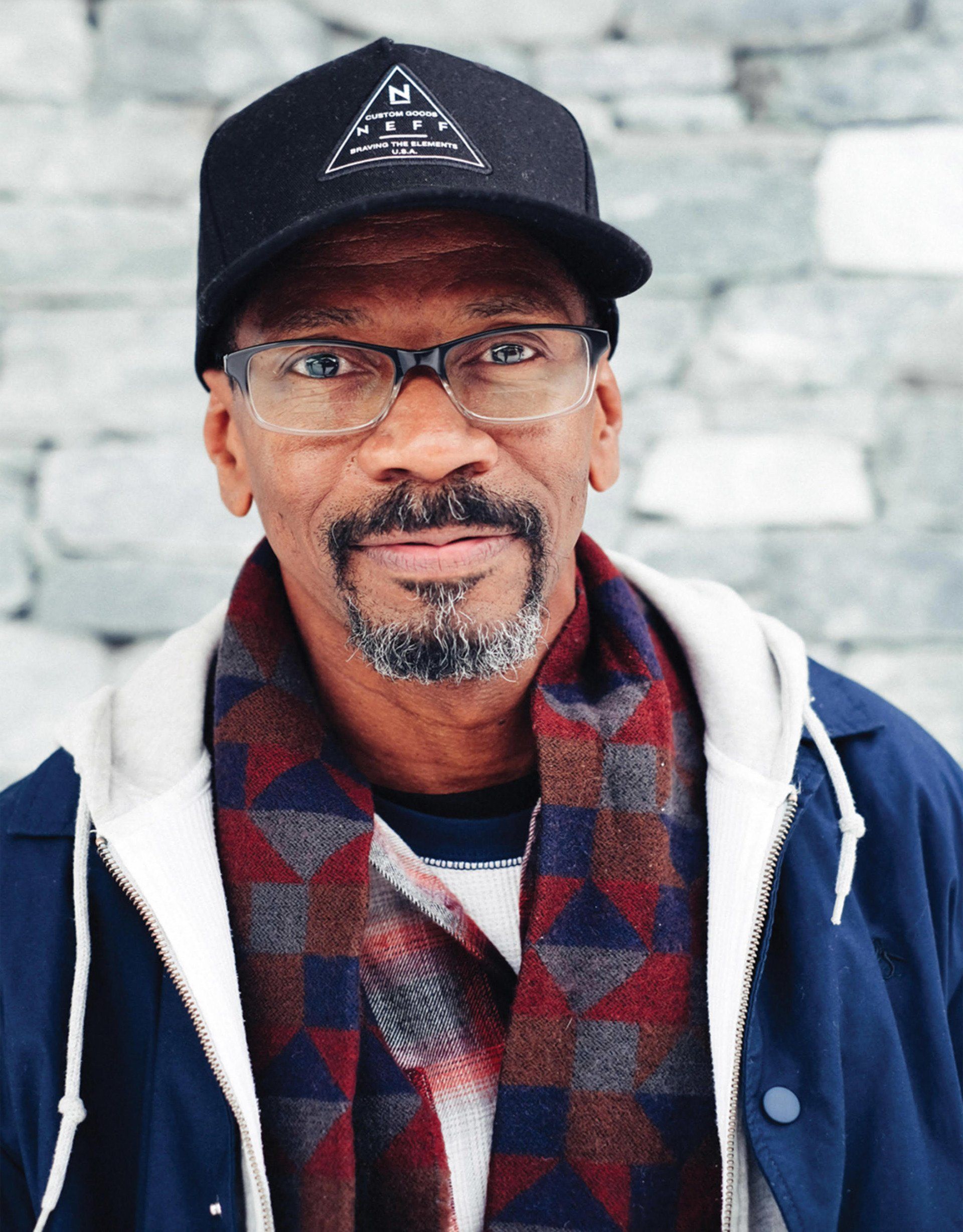 A man wearing a hat and scarf is standing in front of a brick wall.