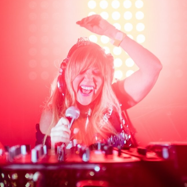 A woman is wearing headphones and holding a microphone in front of a red background.