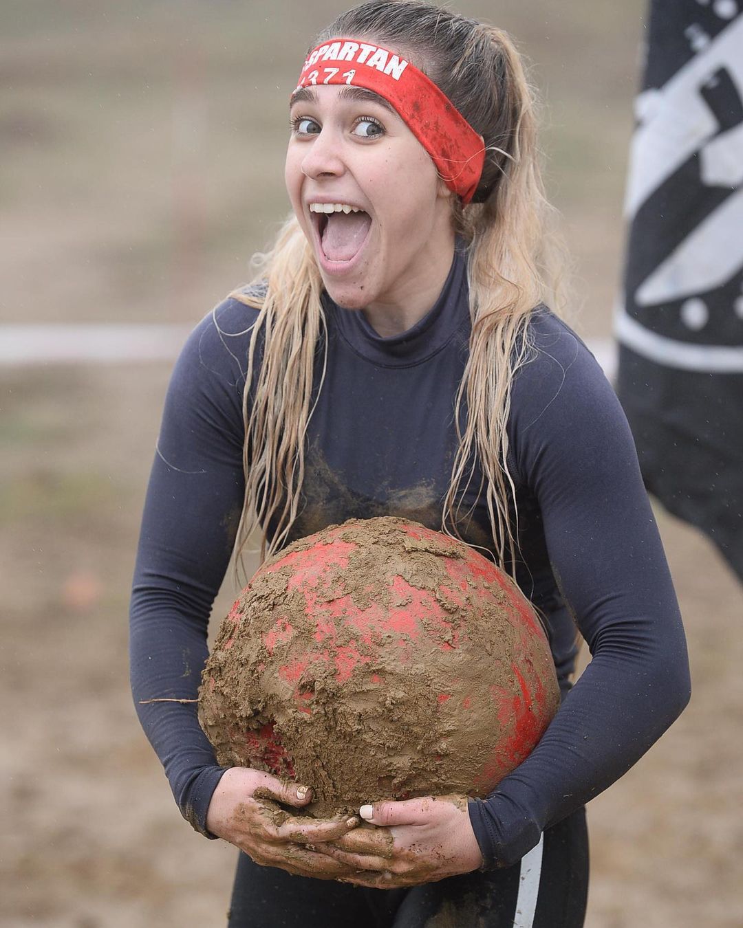 Woman in Spartan Race headband, covered in mud, holding a large, muddy red ball, smiling with mouth open.