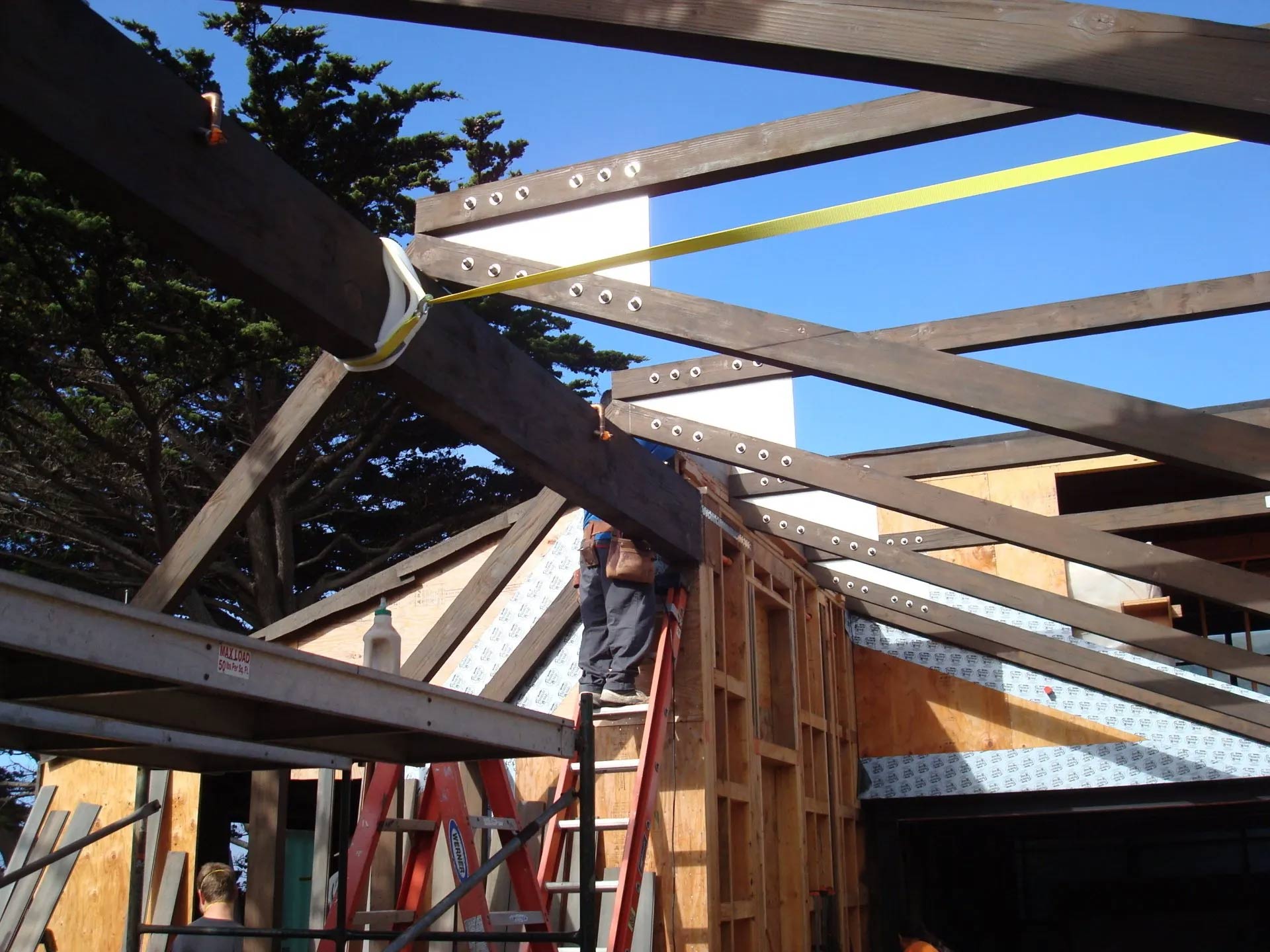 Construction site: Worker on ladder installing wood beams for a roof frame. Yellow strap secures a beam.