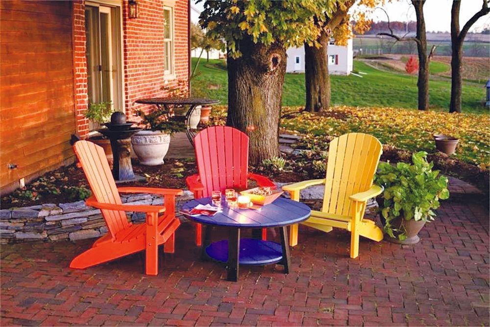 Three colorful Adirondack chairs around a blue table on a brick patio, with a red brick house and trees in the background.
