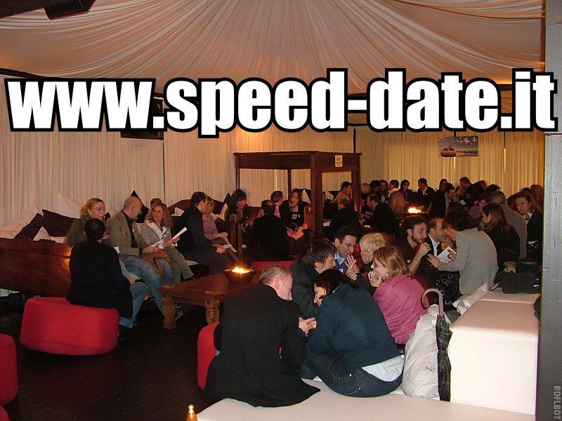 Una stanza scarsamente illuminata con soffitti drappeggiati mostra una folla di persone sedute che partecipano a un evento di speed date.it