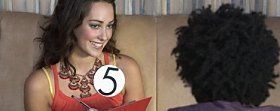 Una donna sorridente, che indossa una maglietta arancione e una grossa collana, tiene in mano un cartello con il numero 5, rivolta verso un uomo