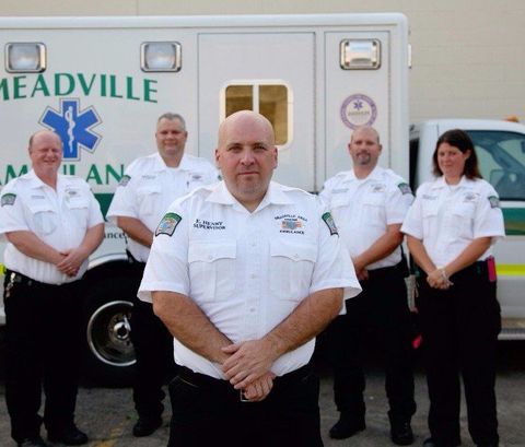 Five ambulance crew members in white uniforms stand in front of a Meadville ambulance, centered on one man